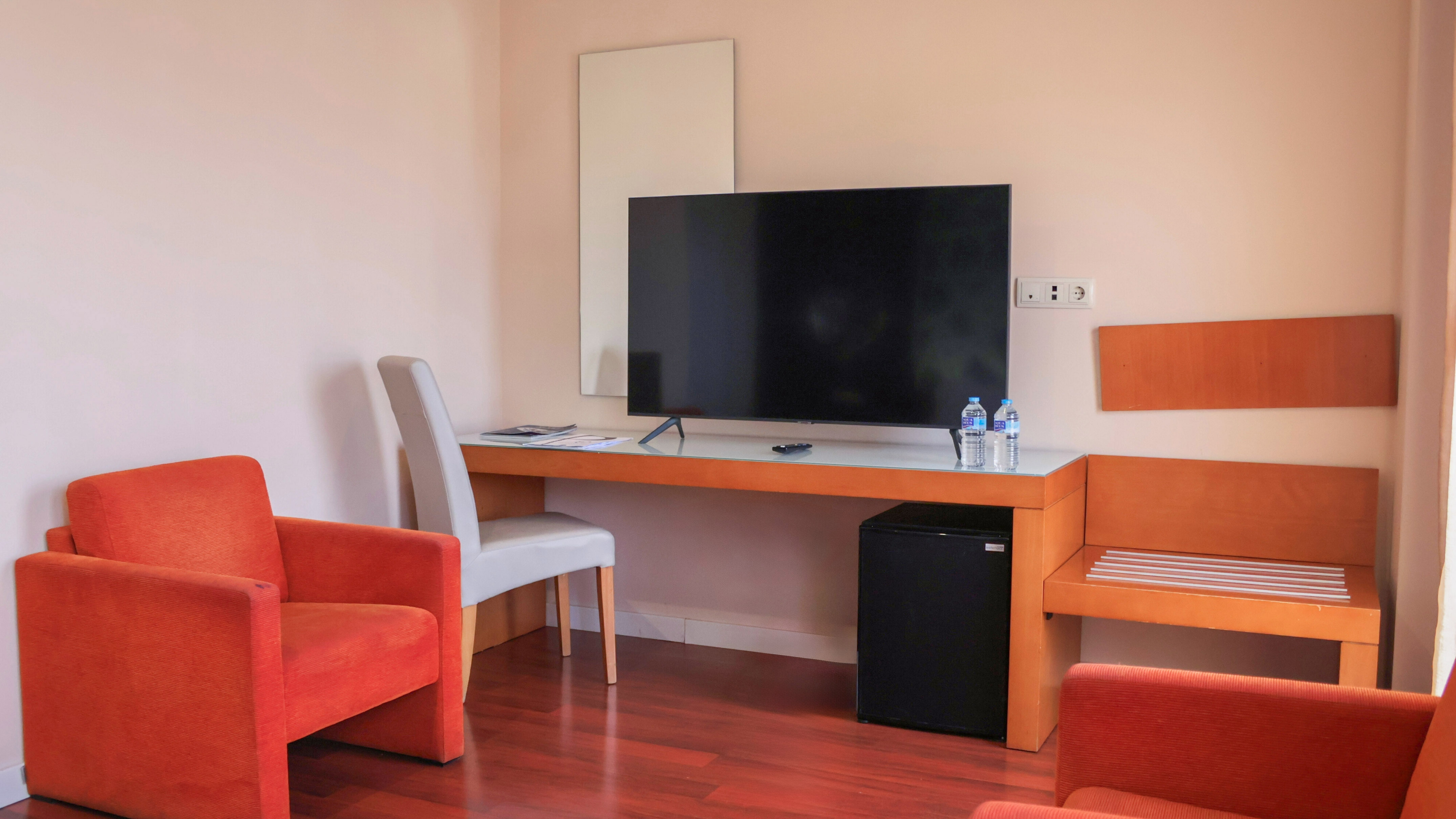 Modern hotel guest room with orange chairs, desk, TV, and minibar.