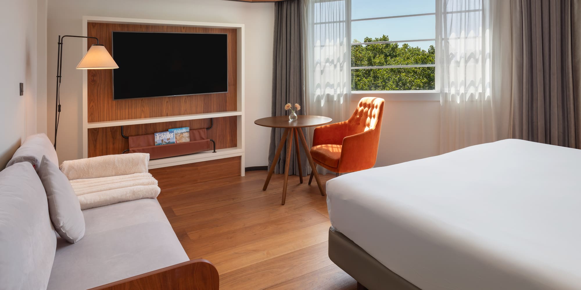 Modern hotel room with bed, sofa, TV, orange chair, and window view.