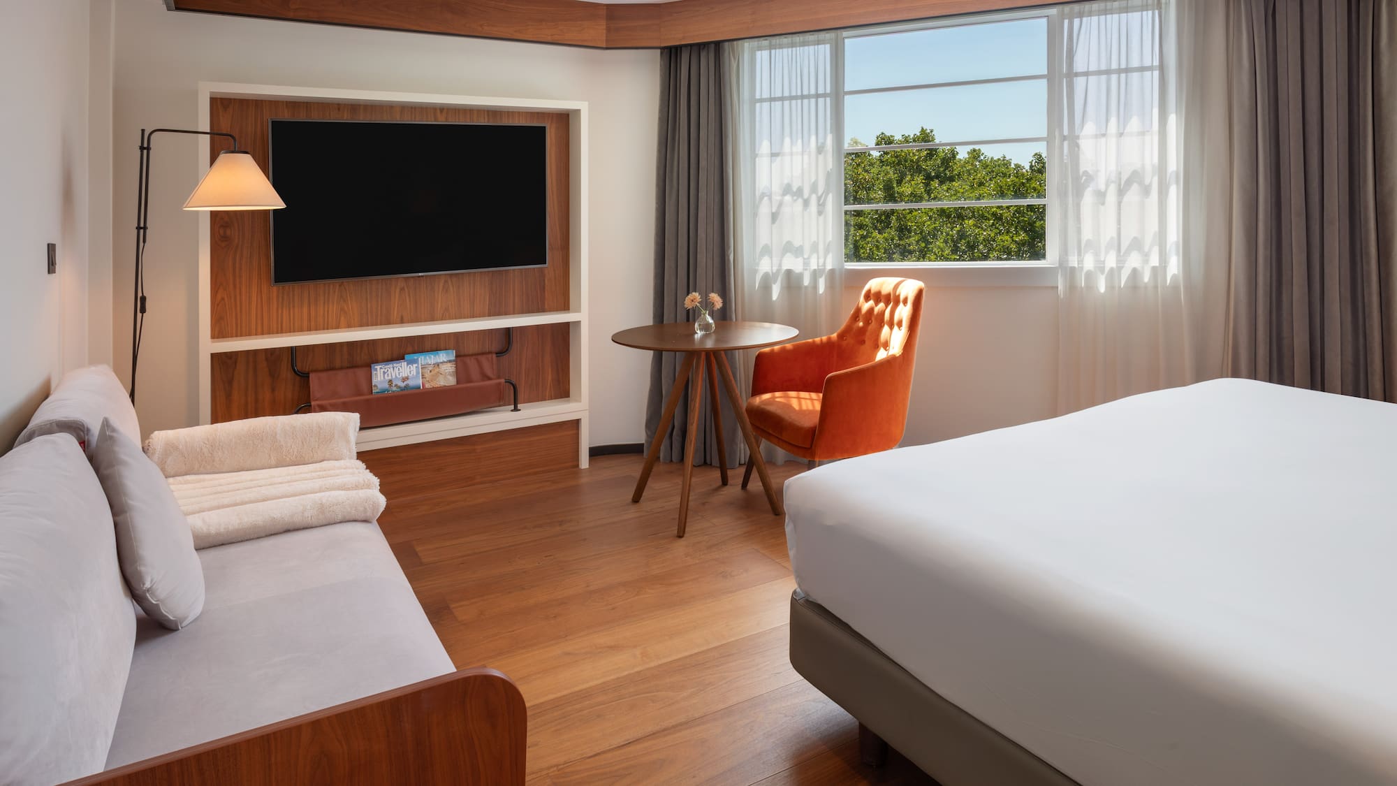 Modern hotel room with bed, sofa, TV, orange armchair, and bright window.