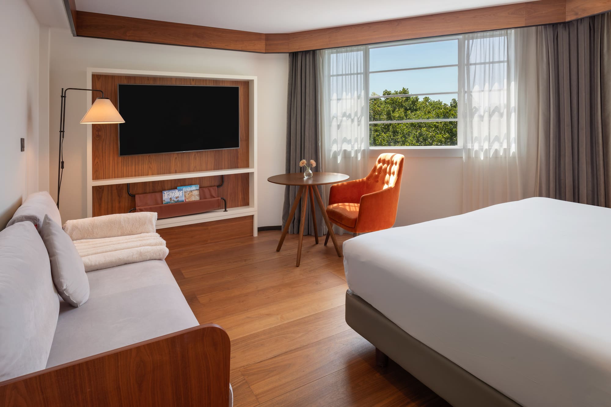 Modern hotel room with bed, sofa, TV, orange armchair, wood floor, and bright window.