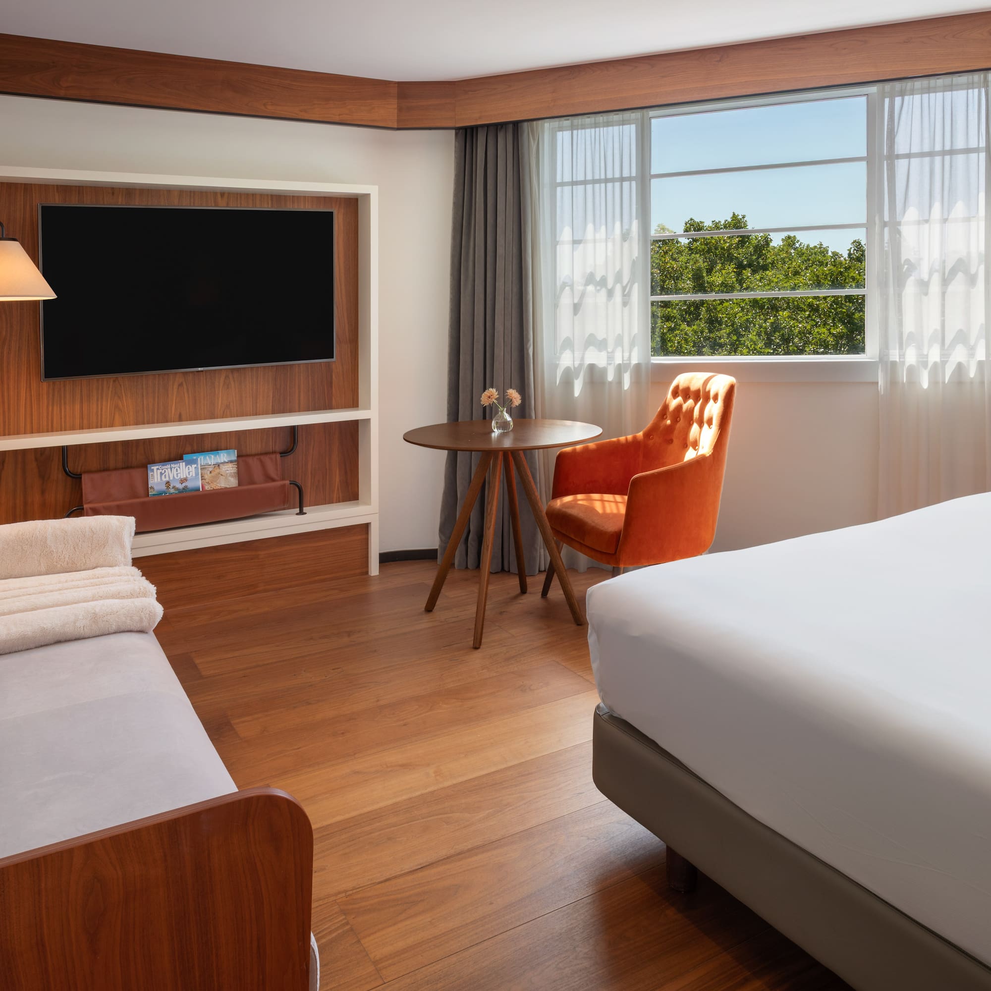 Modern hotel room with bed, sofa, TV, orange armchair, and bright window.