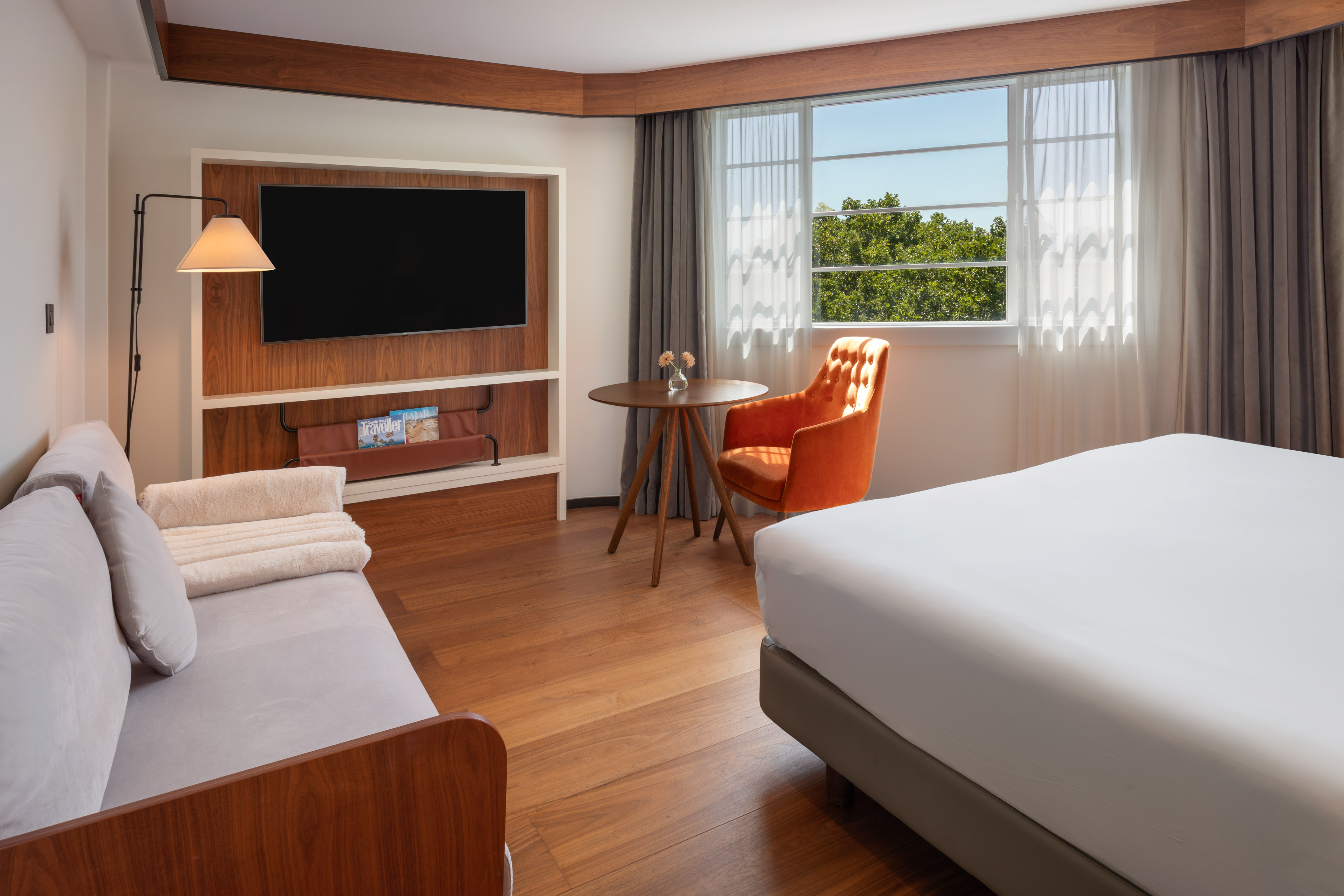 Modern hotel room with bed, sofa, TV, orange armchair, wood floor, and bright window.