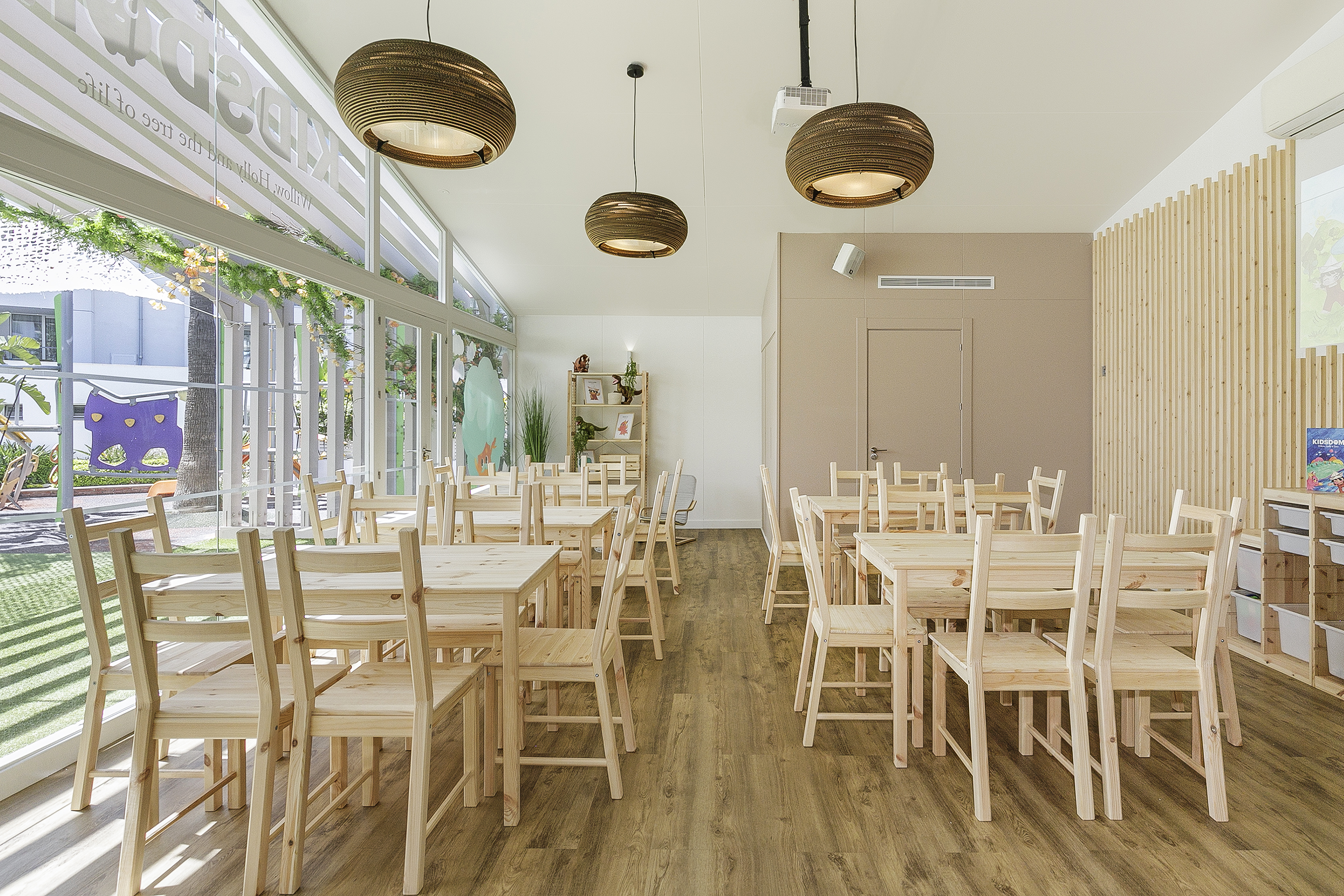 Bright kids classroom with wooden tables, chairs, large windows to play area.