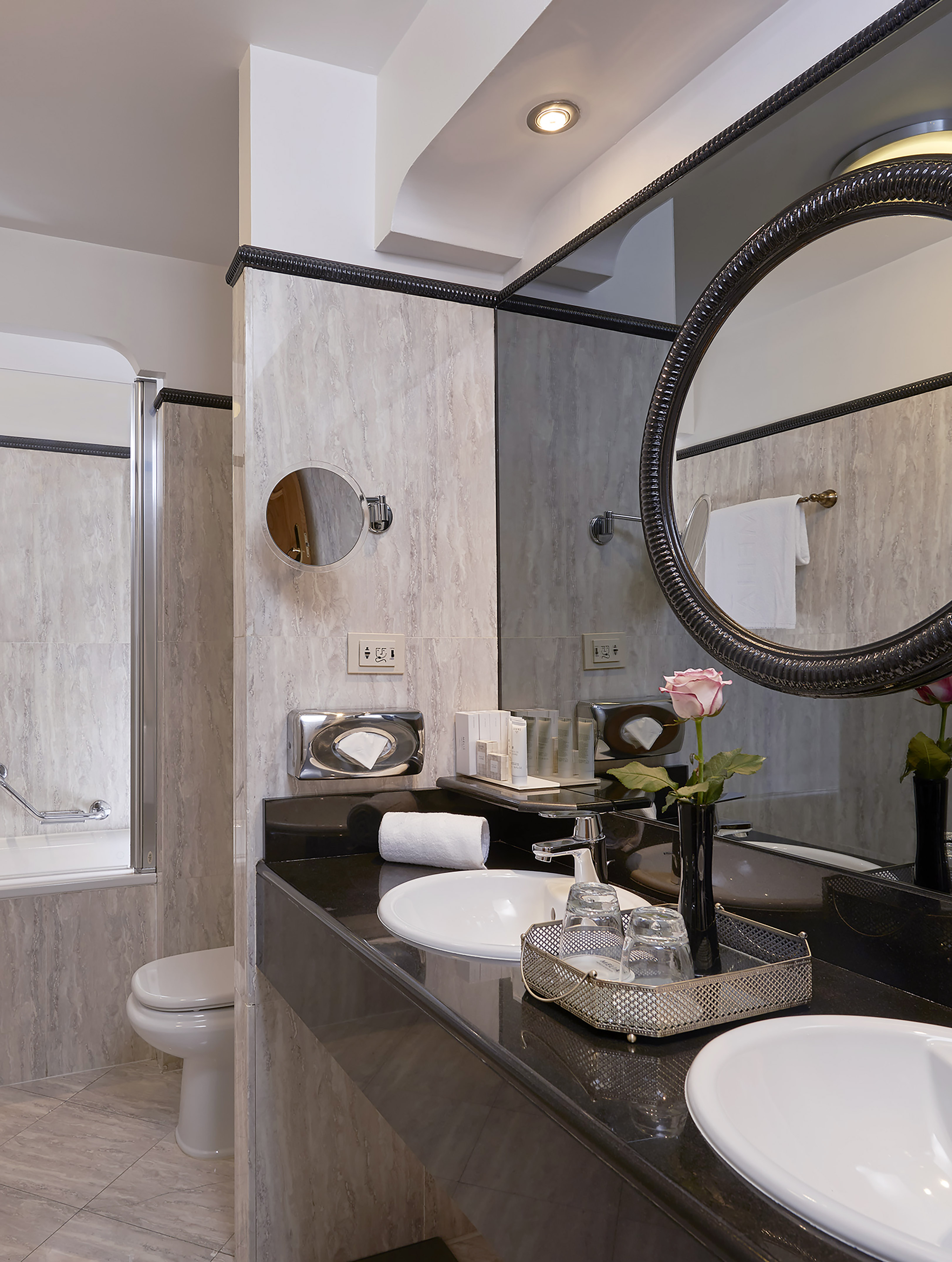 Hotel bathroom with dual sinks, black marble vanity, round mirror, and tub.