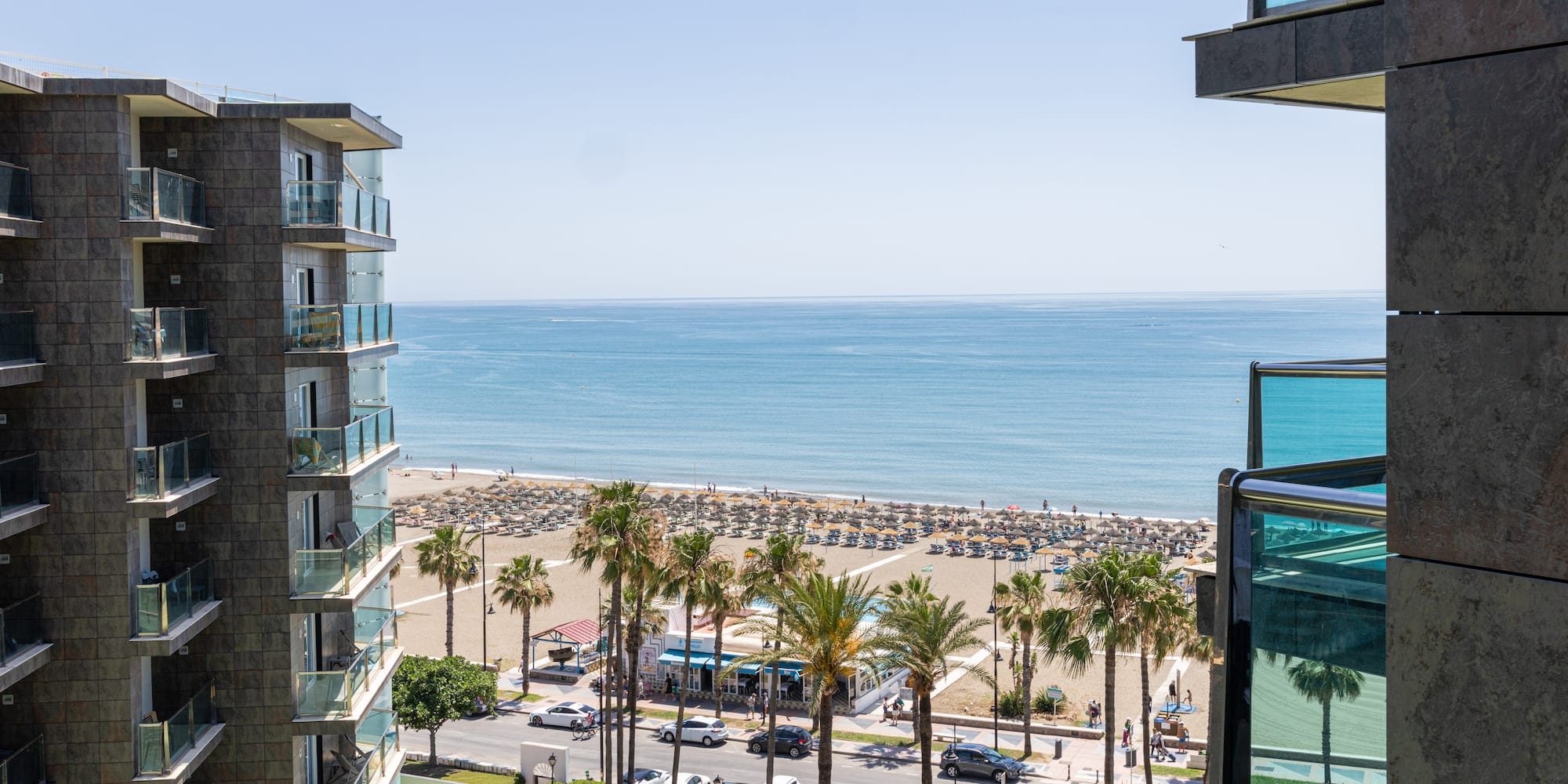 Beachfront hotel view with ocean, sandy beach, palm trees, and building balconies.
