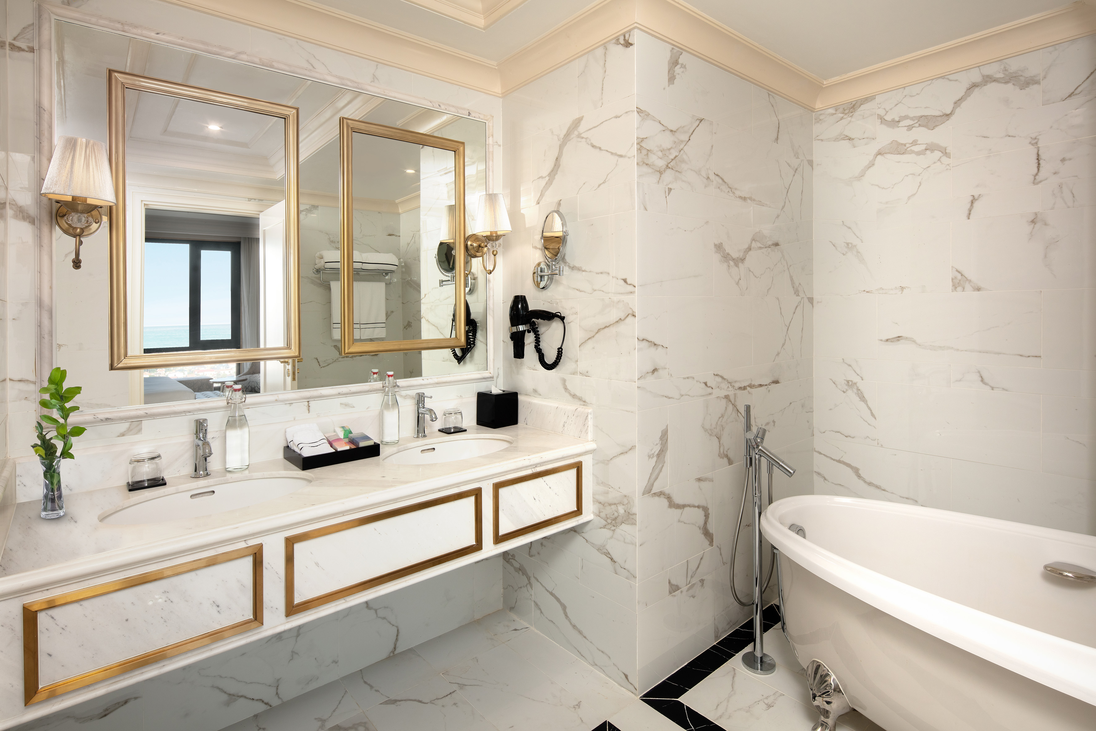 Chic marble bathroom with double vanity, gold mirrors, freestanding tub, and sea view.