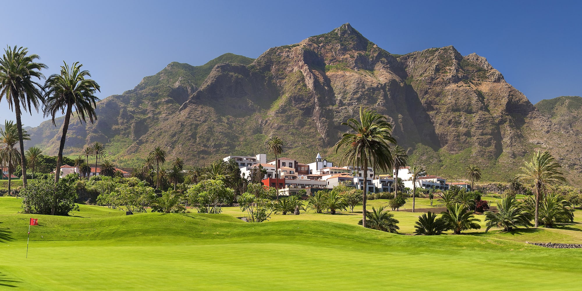 Scenic golf course with palm trees, resort, and rugged mountains.