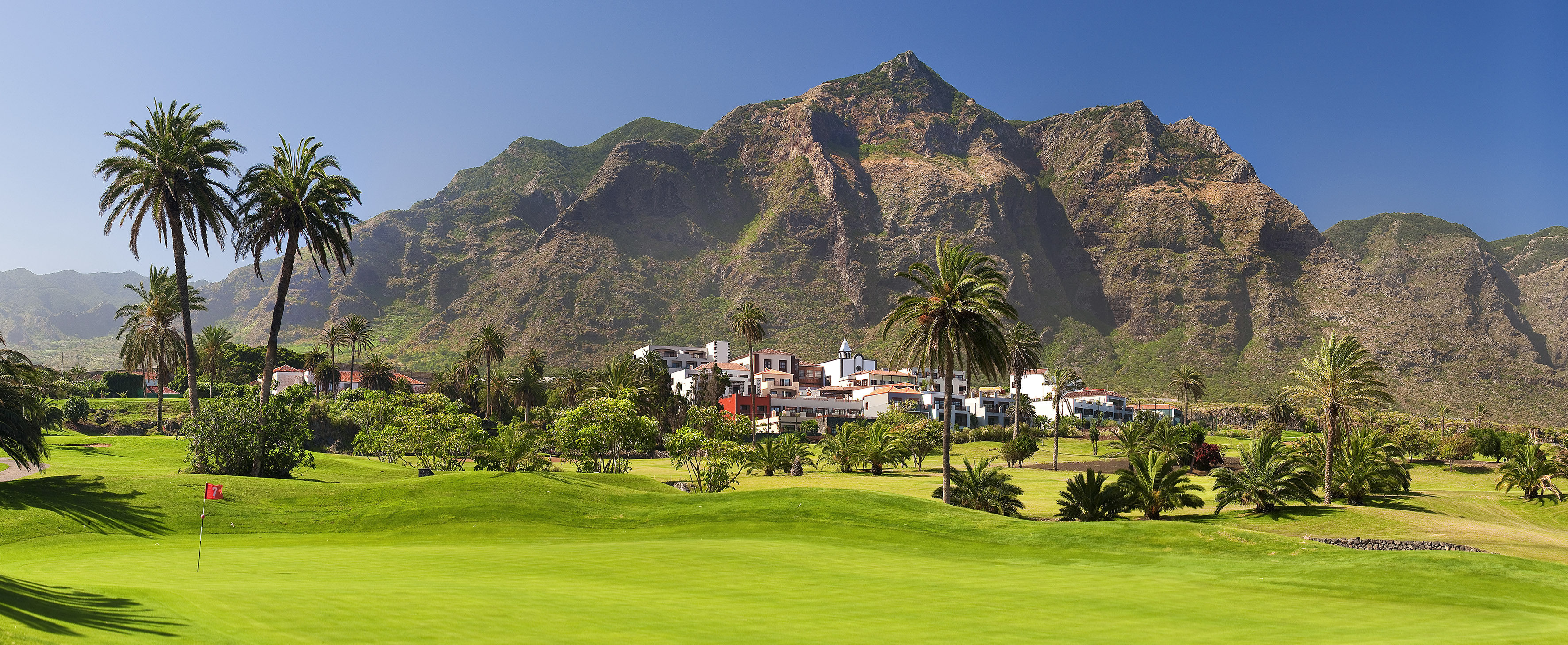Scenic golf course with palm trees, resort, and rugged mountains.