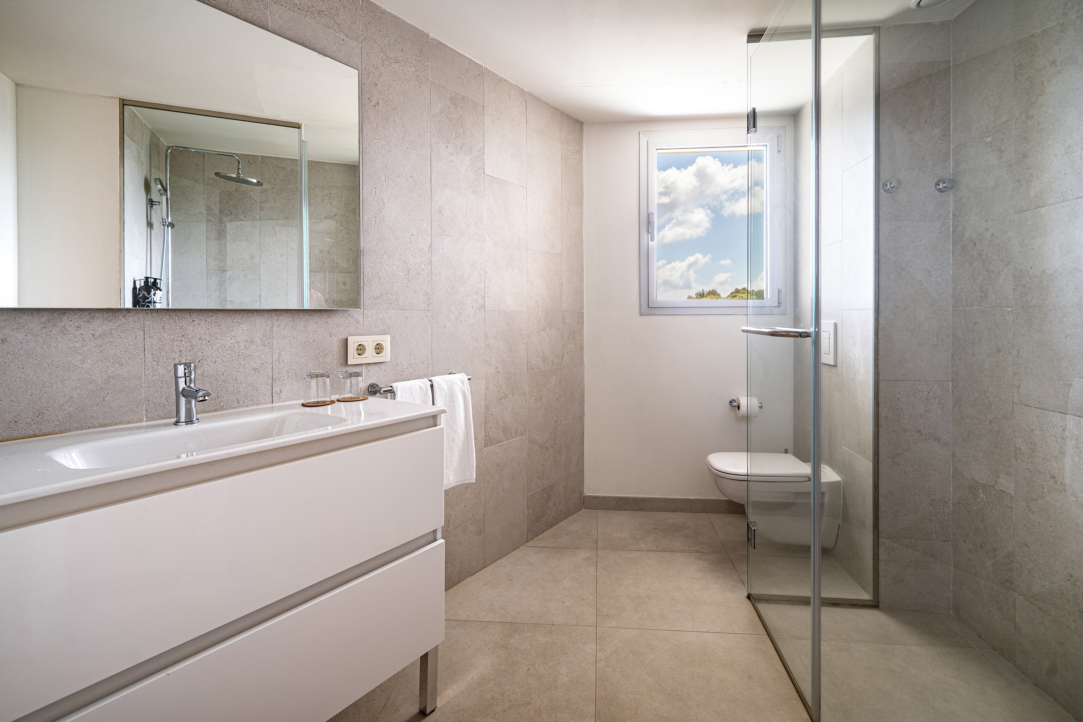 Modern bathroom with white vanity, grey tiled walls, glass shower, toilet, and window.