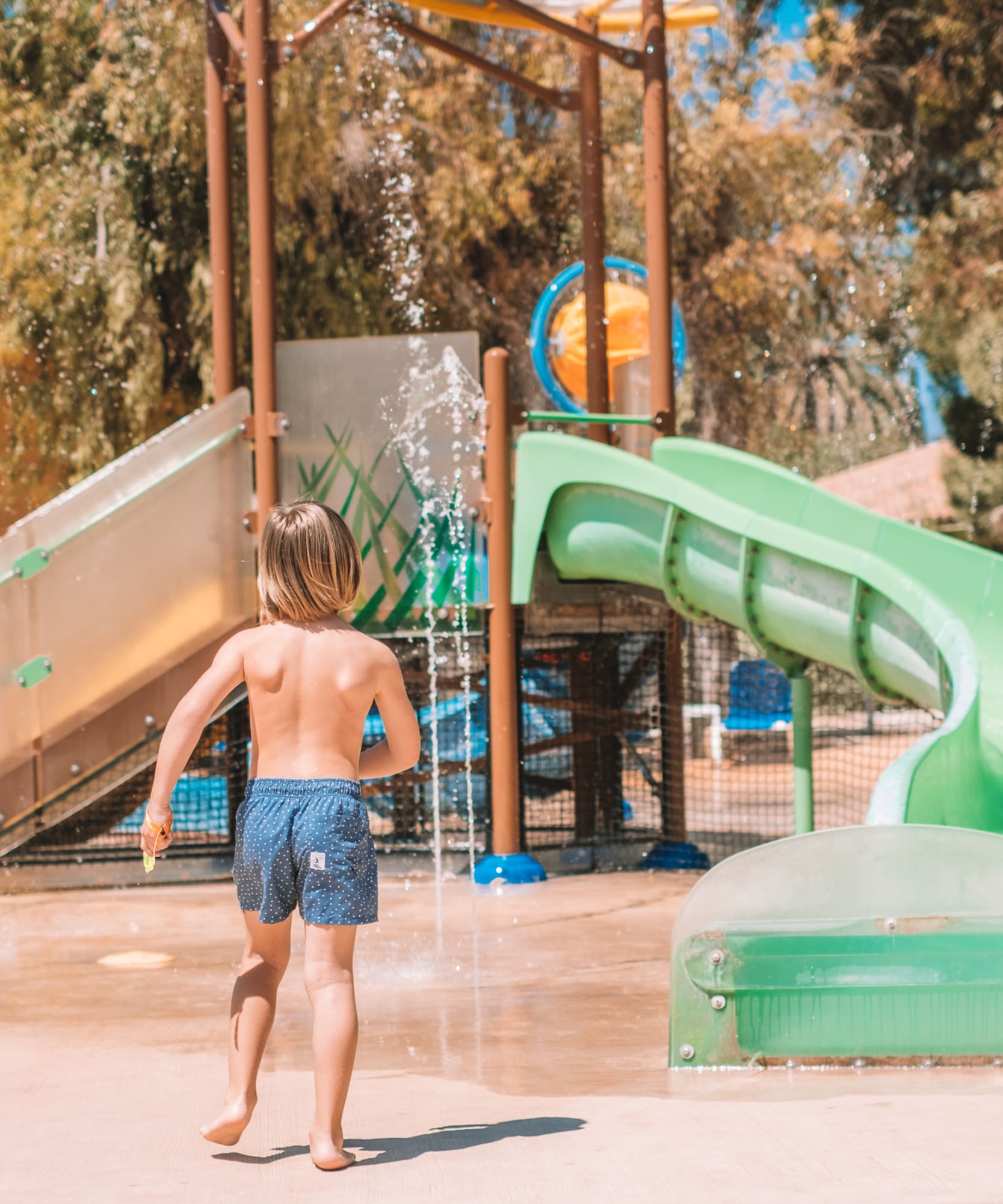 Young boy running towards water slides and splash pad at a sunny water park.