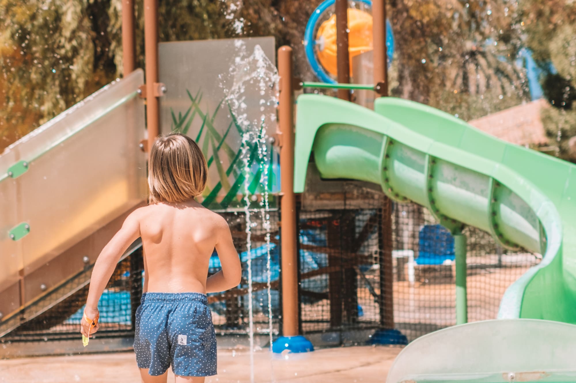 Young boy running towards water slides and splash pad at a sunny water park.