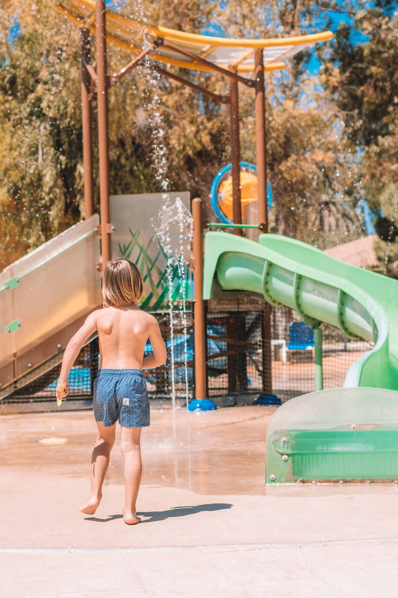 Young boy running towards water slides and splash pad at a sunny water park.