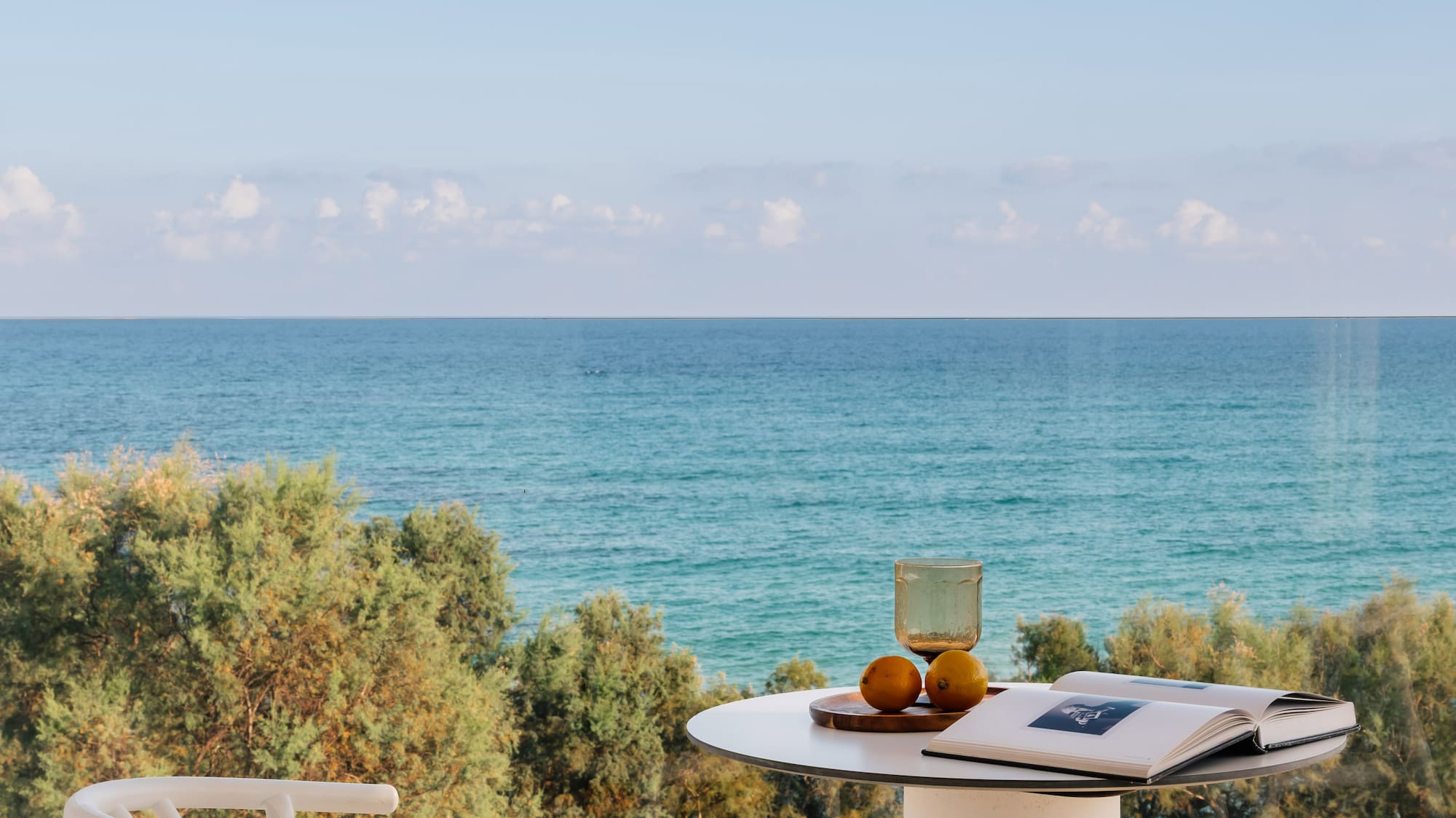 Hotel balcony with ocean view, white table, chairs, book, and fruit.