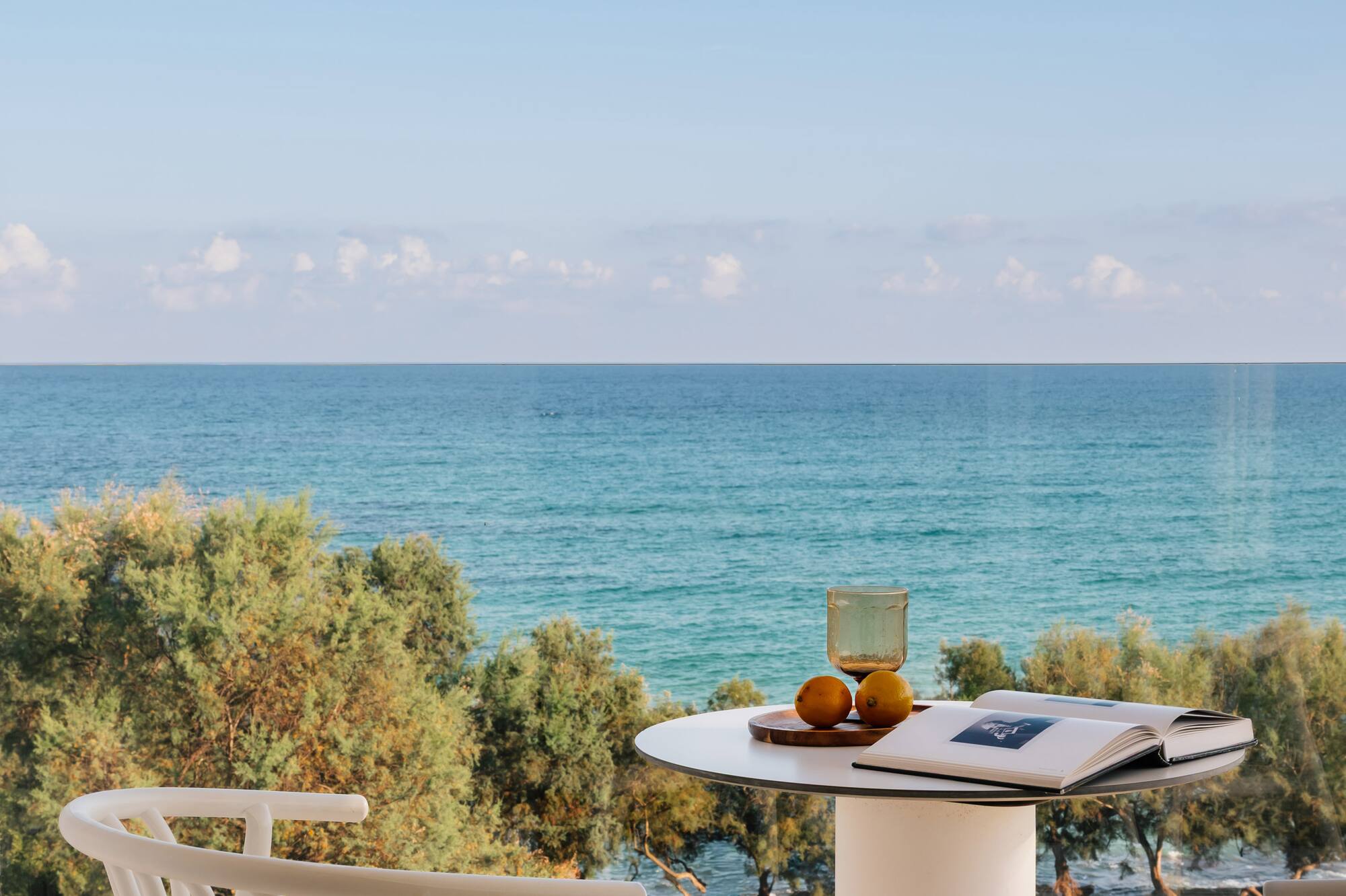 Balcón con vista al mar, mesa blanca con libro y naranjas. Relax costero.