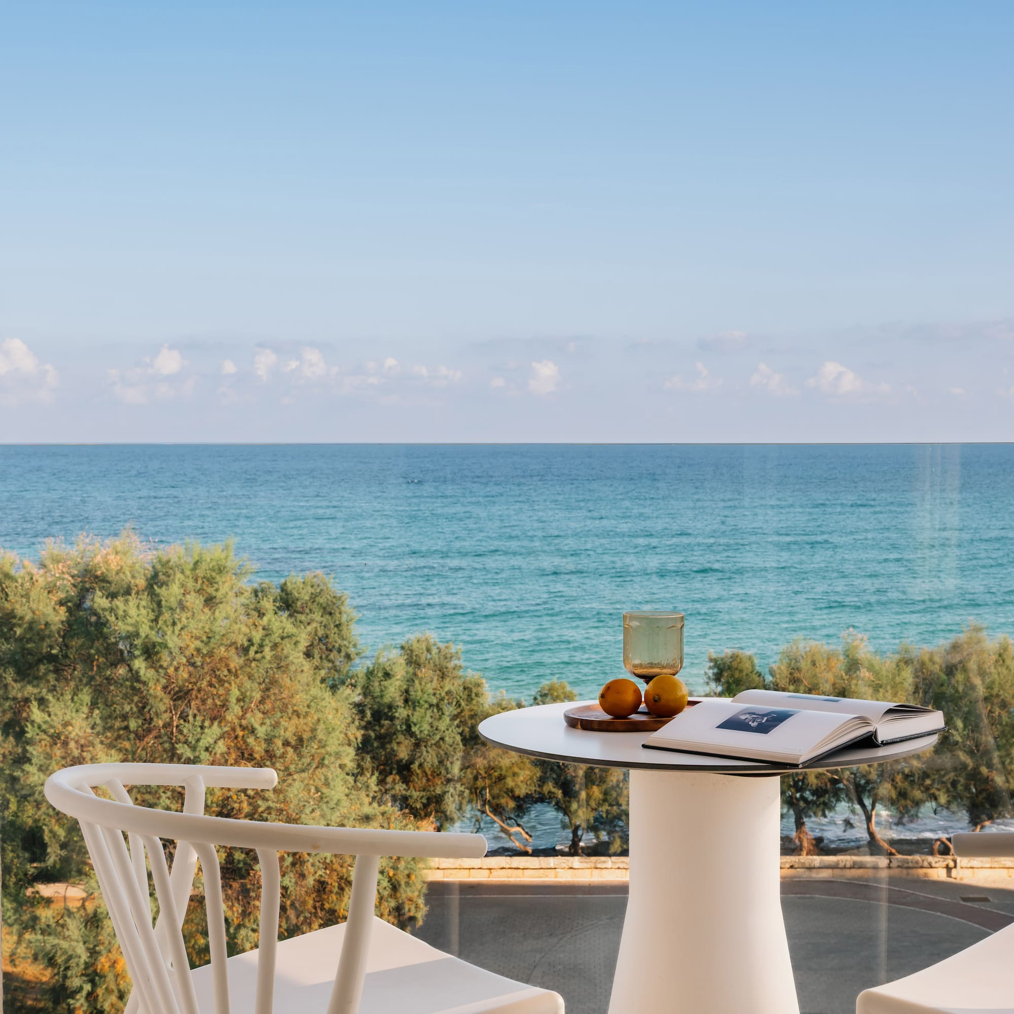 Hotel balcony with ocean view, white table, chairs, book, and fruit.
