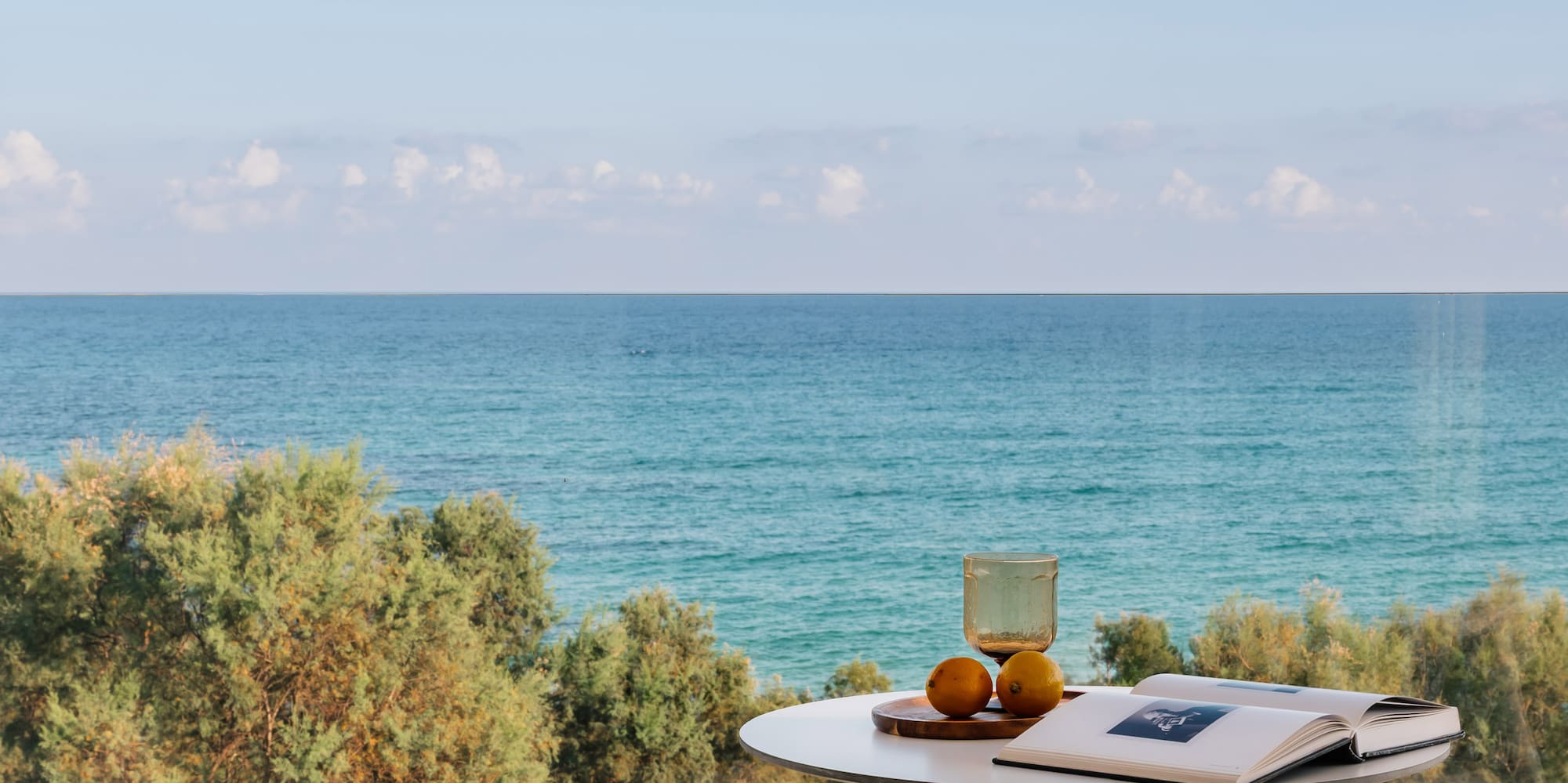 Hotel balcony with ocean view, white table, chairs, book, and fruit.