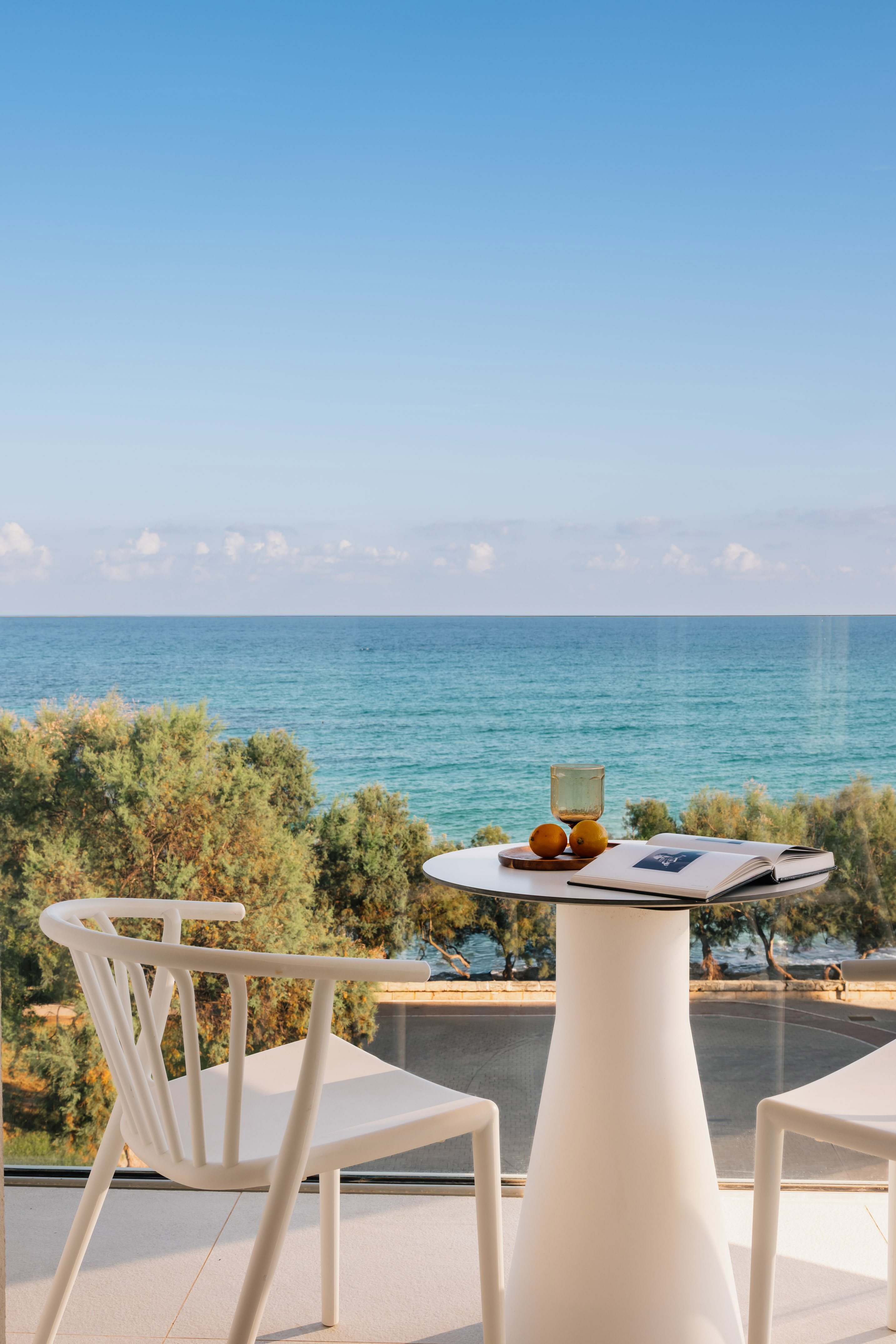 Hotel balcony with ocean view, white table, chairs, book, and fruit.