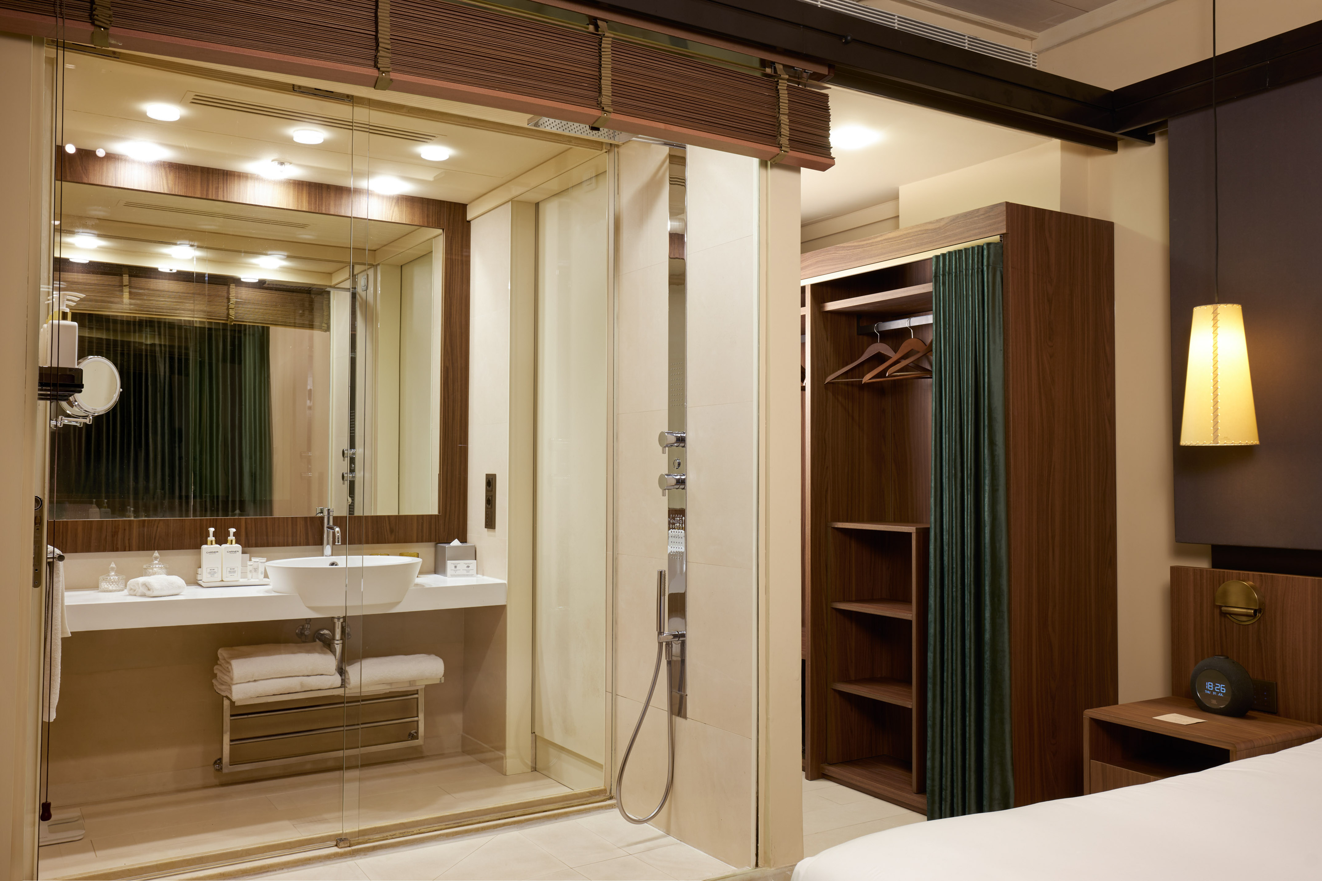 Modern hotel room: glass bathroom, vanity, shower, open wooden closet.