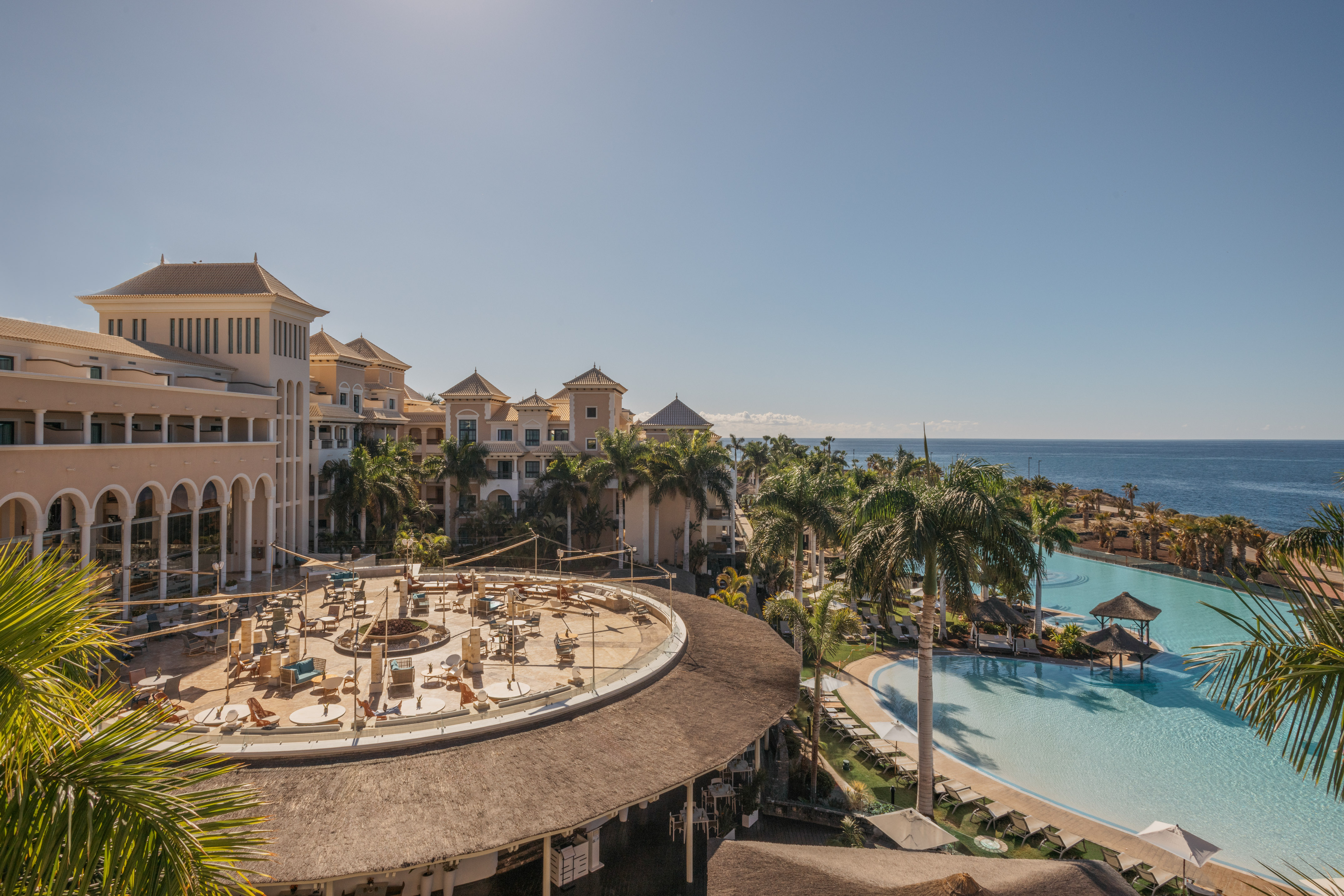Luxury resort with ocean, large swimming pool, palm trees, and outdoor terrace.