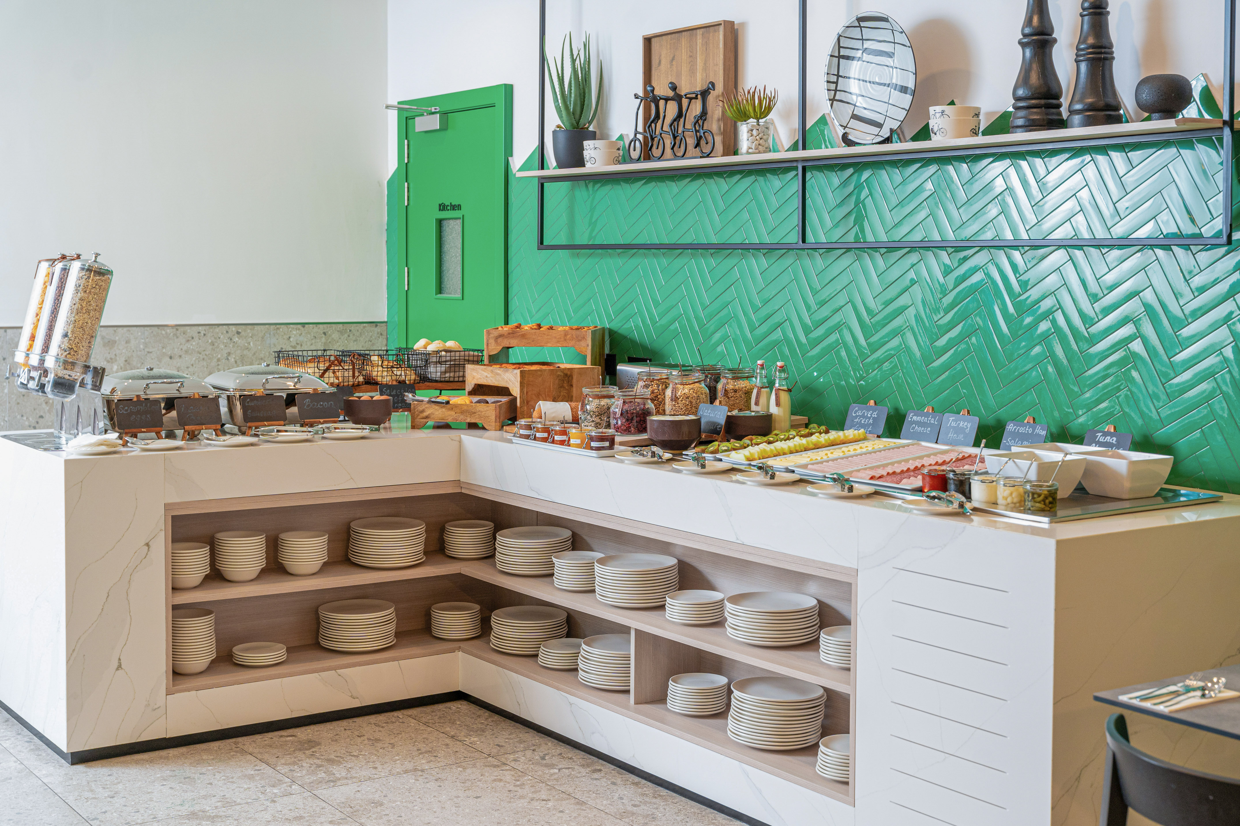 Breakfast buffet with hot food, cold cuts, pastries, cereals, green tiled wall.