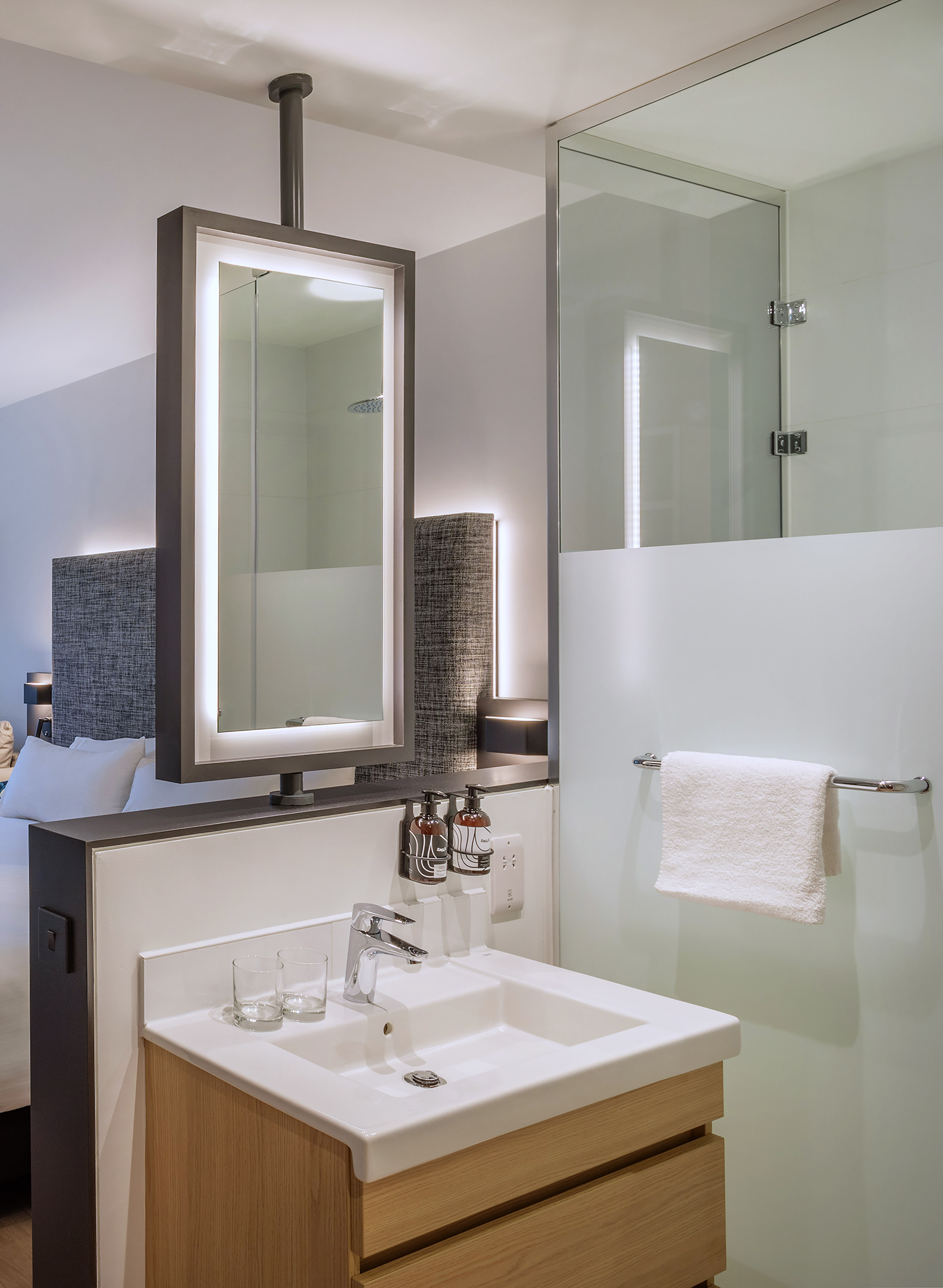 Modern hotel bathroom with wood vanity, backlit mirror, and glass shower.