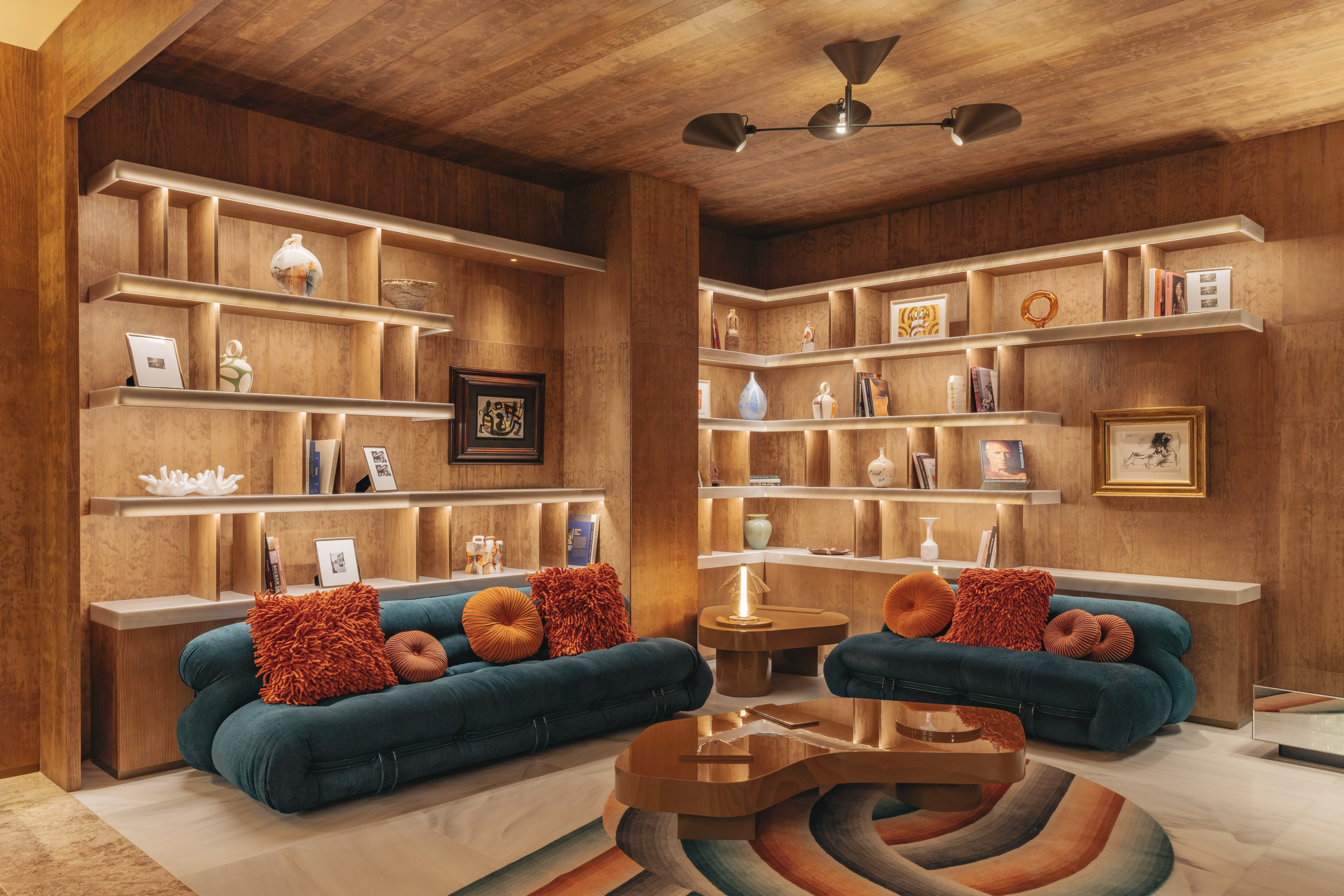 Modern lounge with teal sofas, orange cushions, lit wooden shelves, and art.