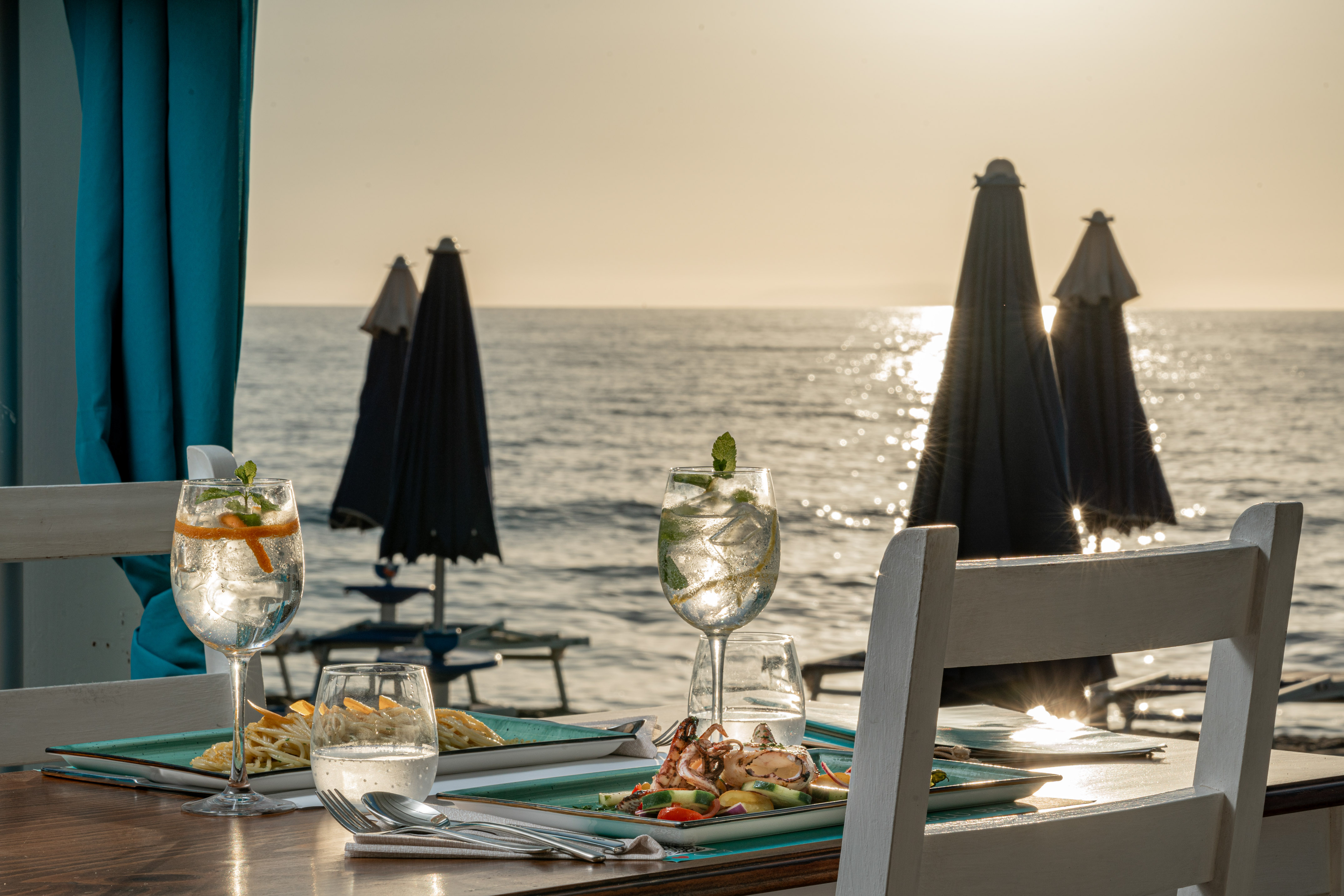 Beachfront restaurant table with cocktails, seafood, and ocean sunset view.