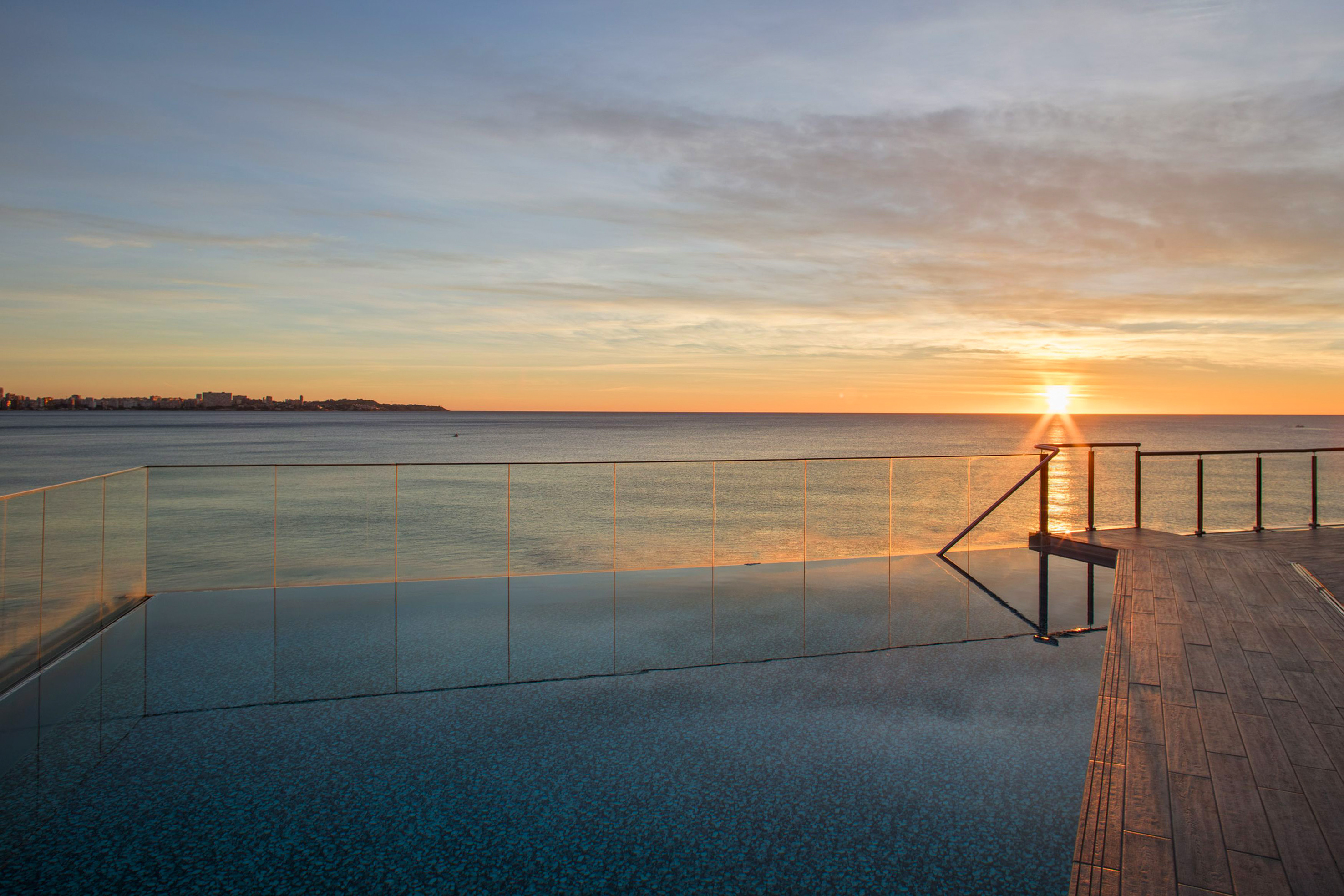 Sunset ocean view from infinity pool, with wooden deck and city skyline.