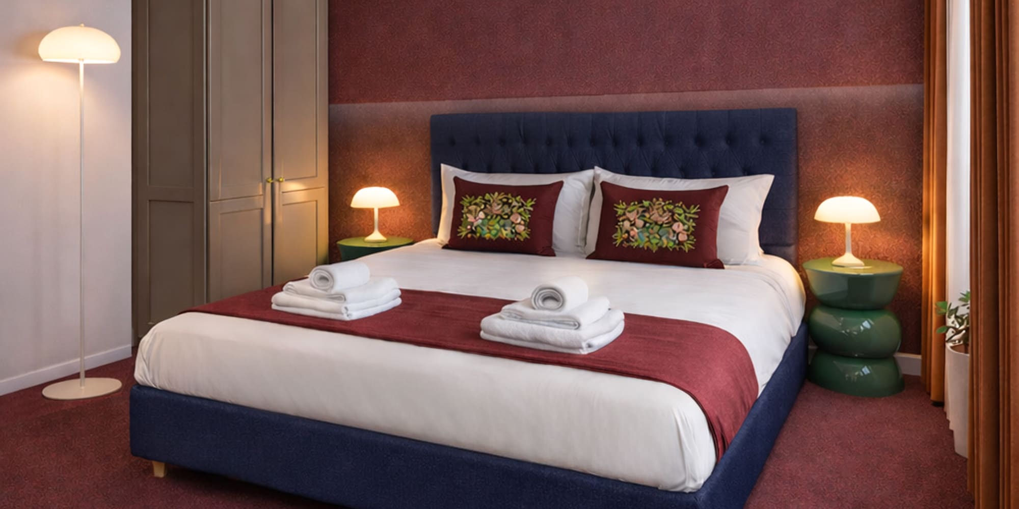 Modern hotel bedroom with navy blue bed, red accent wall, and green nightstands.