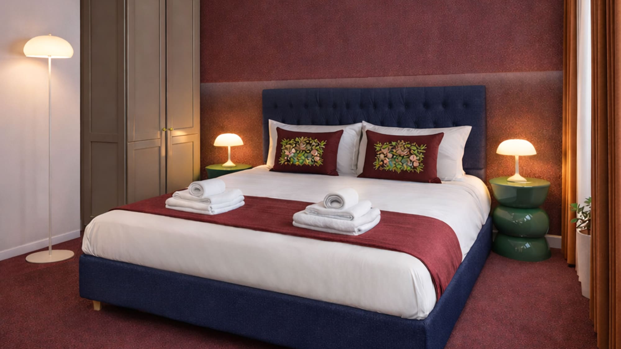 Modern bedroom with blue upholstered bed, red bedding, green nightstands, soft light.