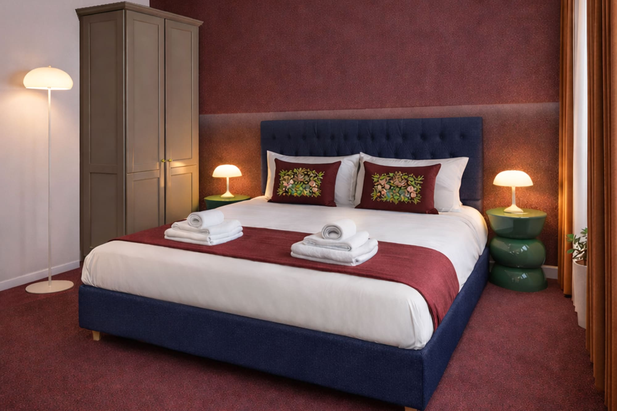 Modern hotel bedroom with navy blue bed, red accent wall, and green nightstands.
