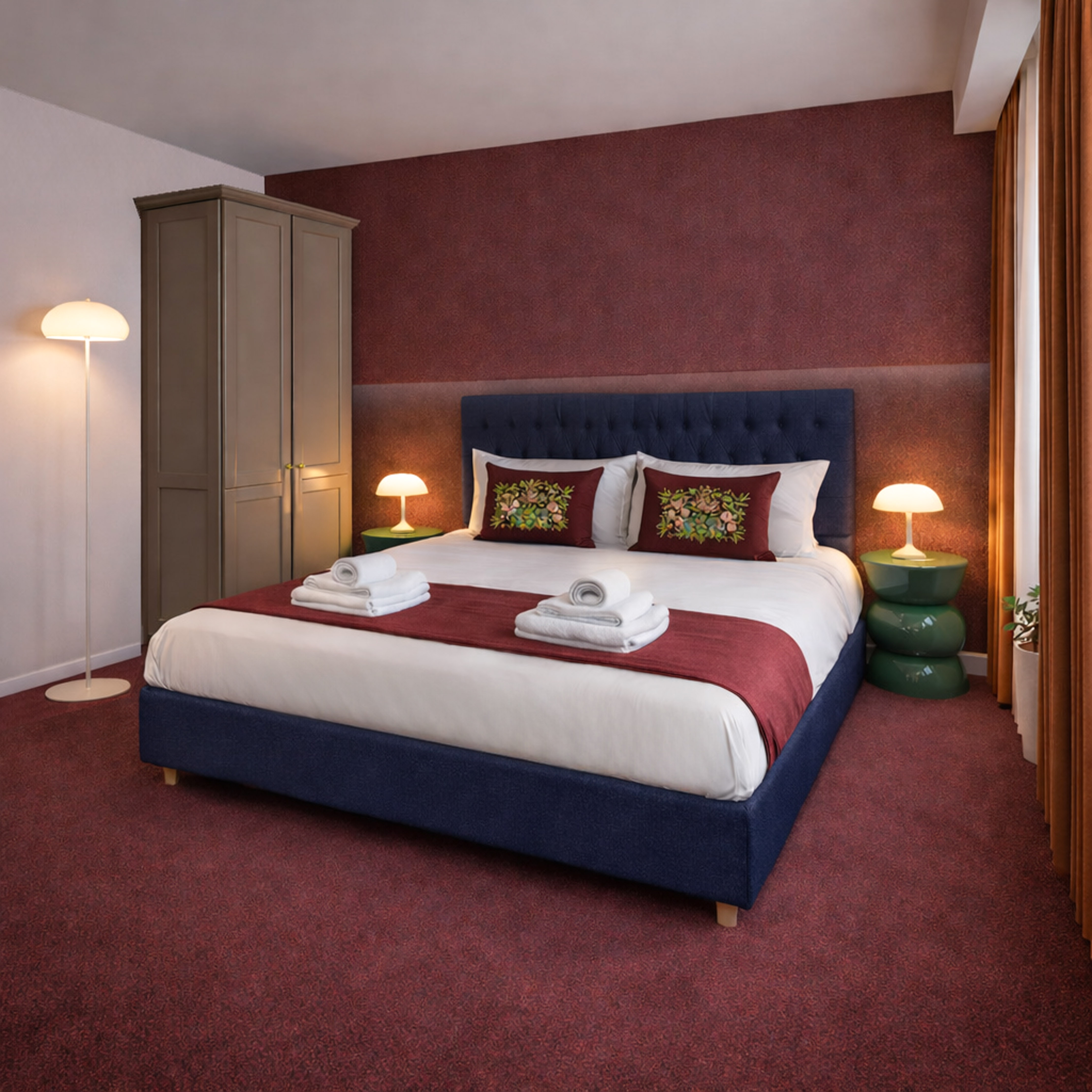 Modern hotel bedroom with navy blue bed, red accent wall, and green nightstands.