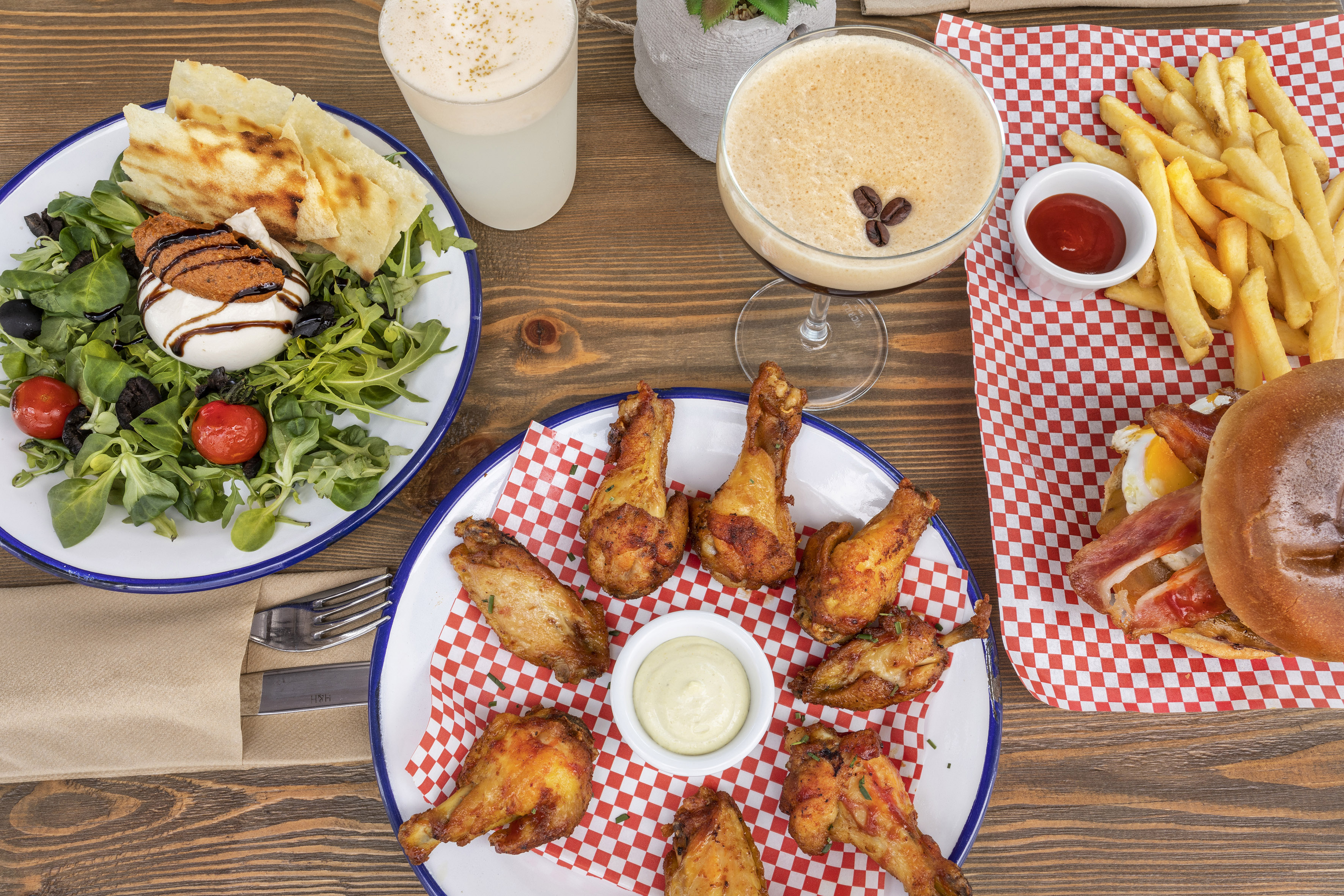Burrata salad, fried chicken, burger, french fries, and cocktails on a wooden table.