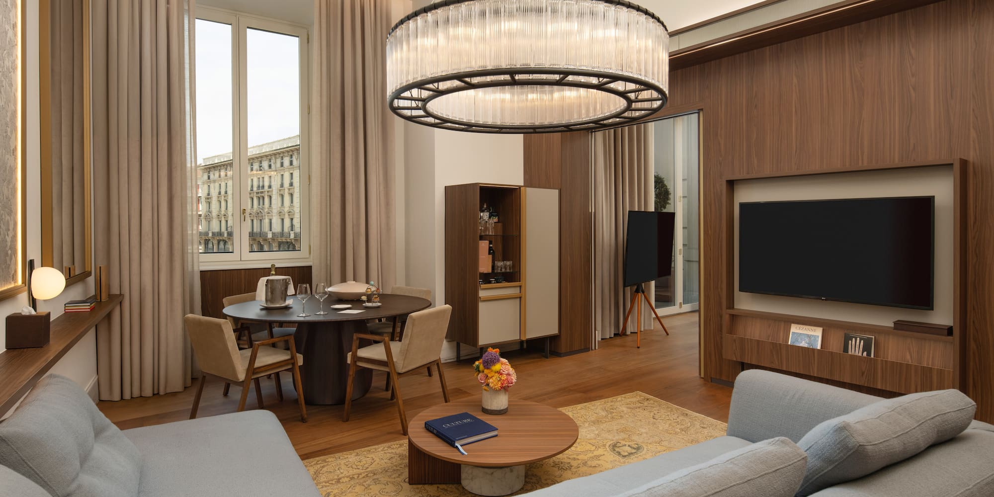 Modern luxury suite with grey sofa, dining area, city view, and elegant chandelier.