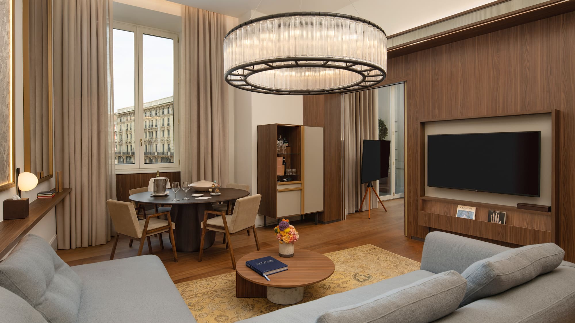 Modern luxury suite with grey sofa, dining area, city view, and elegant chandelier.