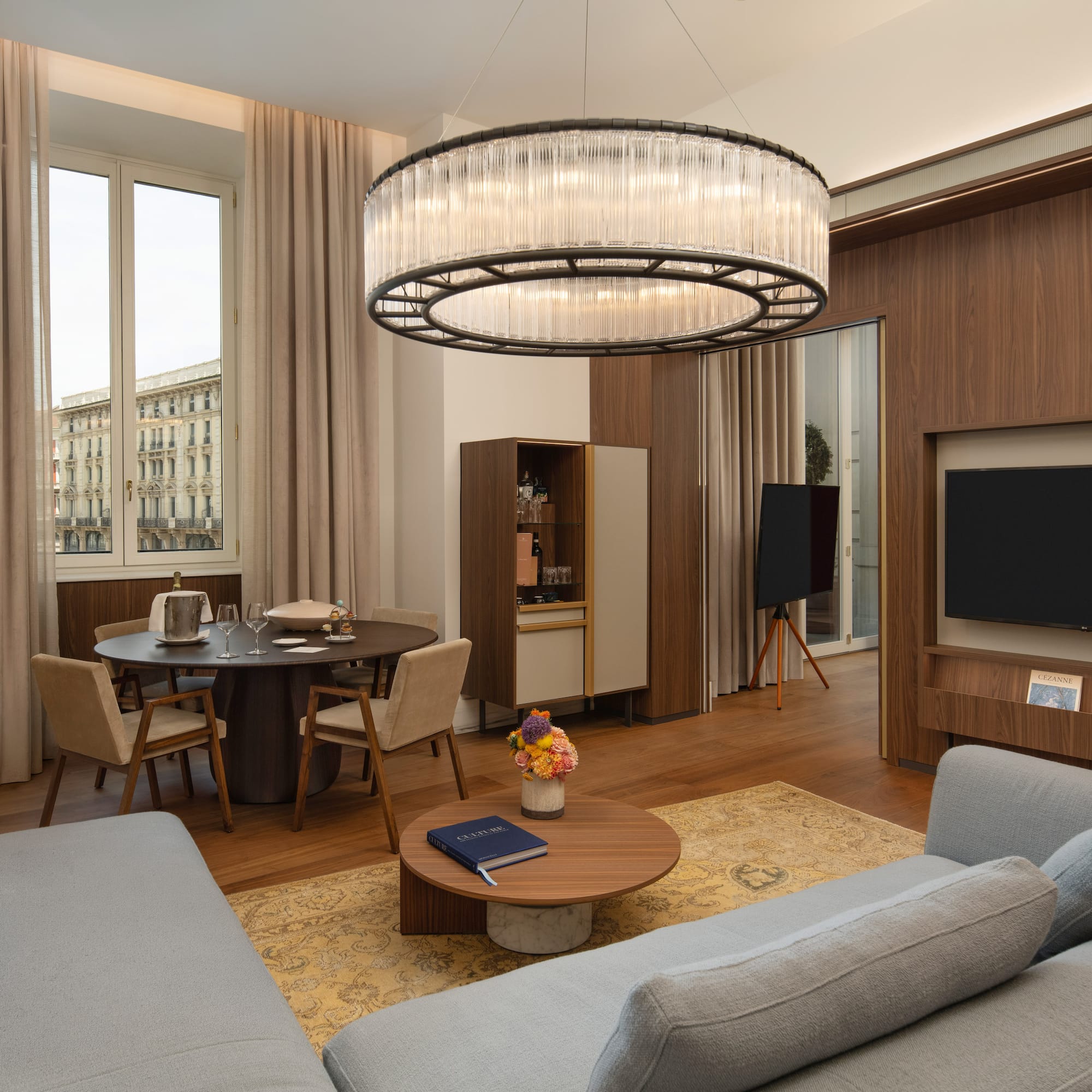 Modern luxury suite with grey sofa, dining area, city view, and elegant chandelier.