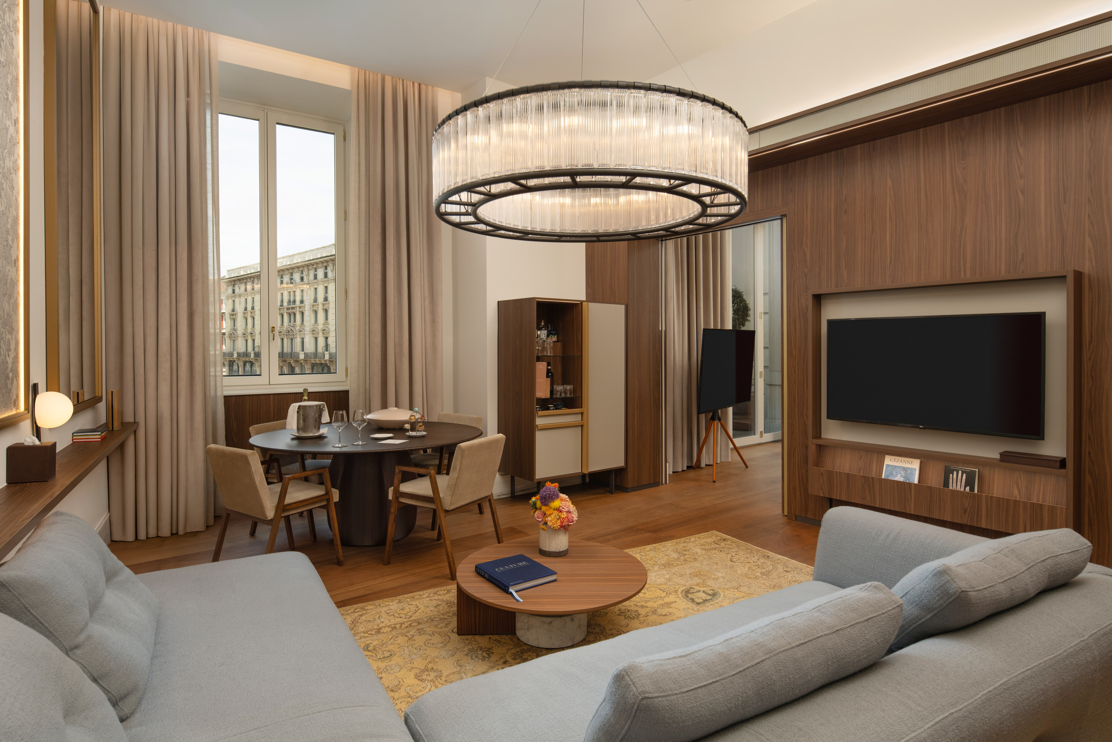 Modern luxury suite with grey sofa, dining area, city view, and elegant chandelier.