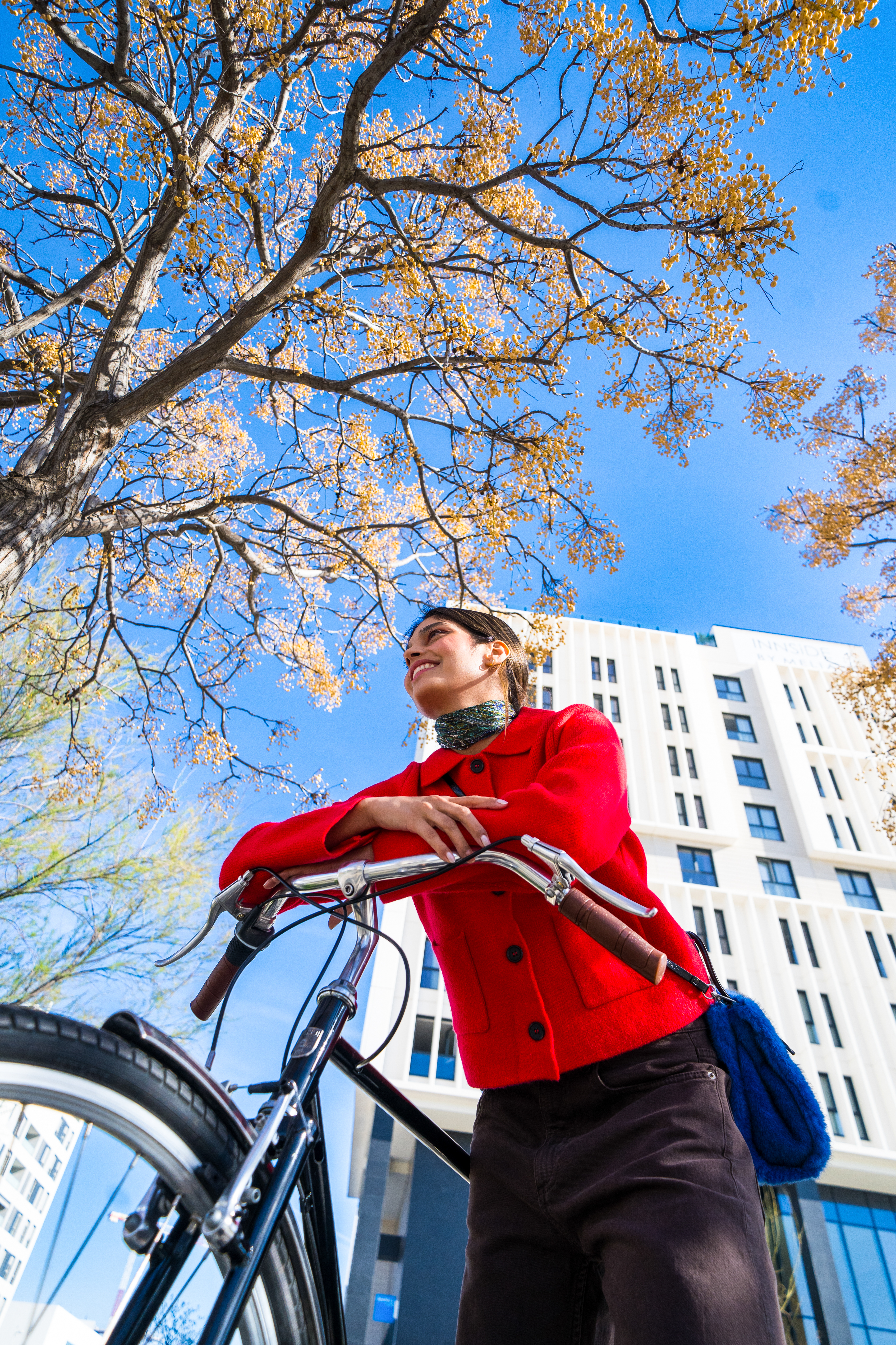 Smiling woman in red jacket with bicycle under autumn tree, city backdrop.