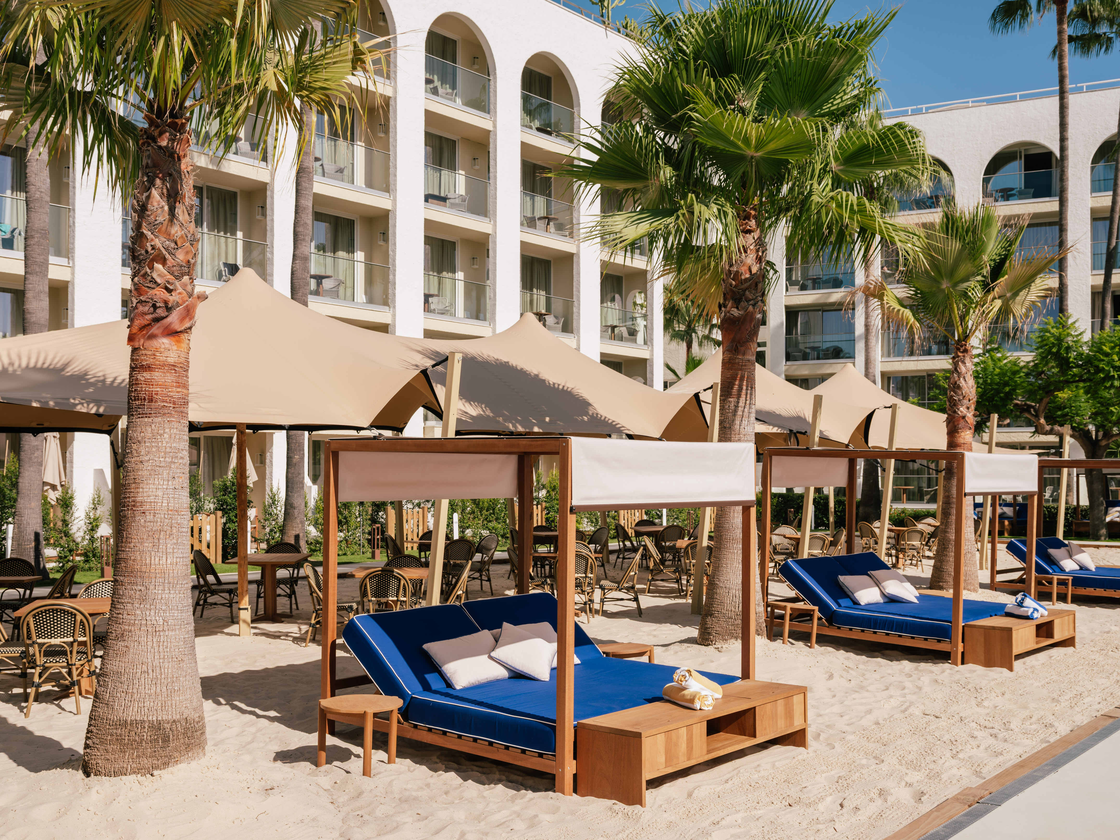 Luxury resort with palm trees, sandy beach lounge, cabanas, and blue daybeds.