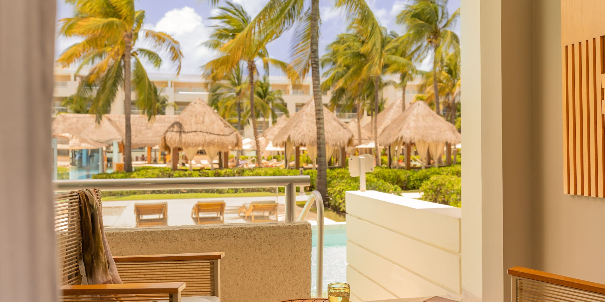 Tropical resort balcony overlooking pool with palm trees and cabanas.