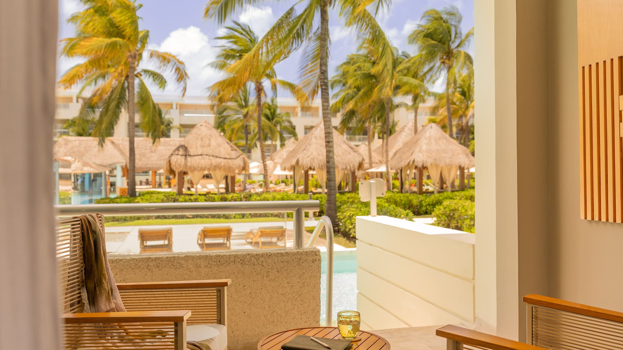 Tropical resort balcony overlooking pool with palm trees and cabanas.