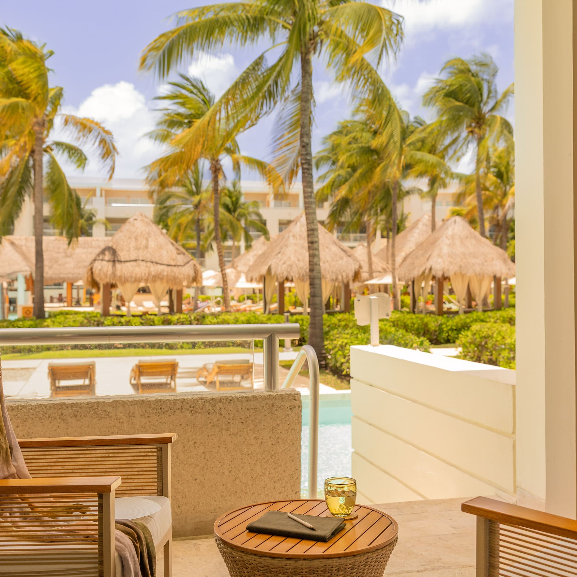 Tropical resort balcony overlooking pool with palm trees and cabanas.