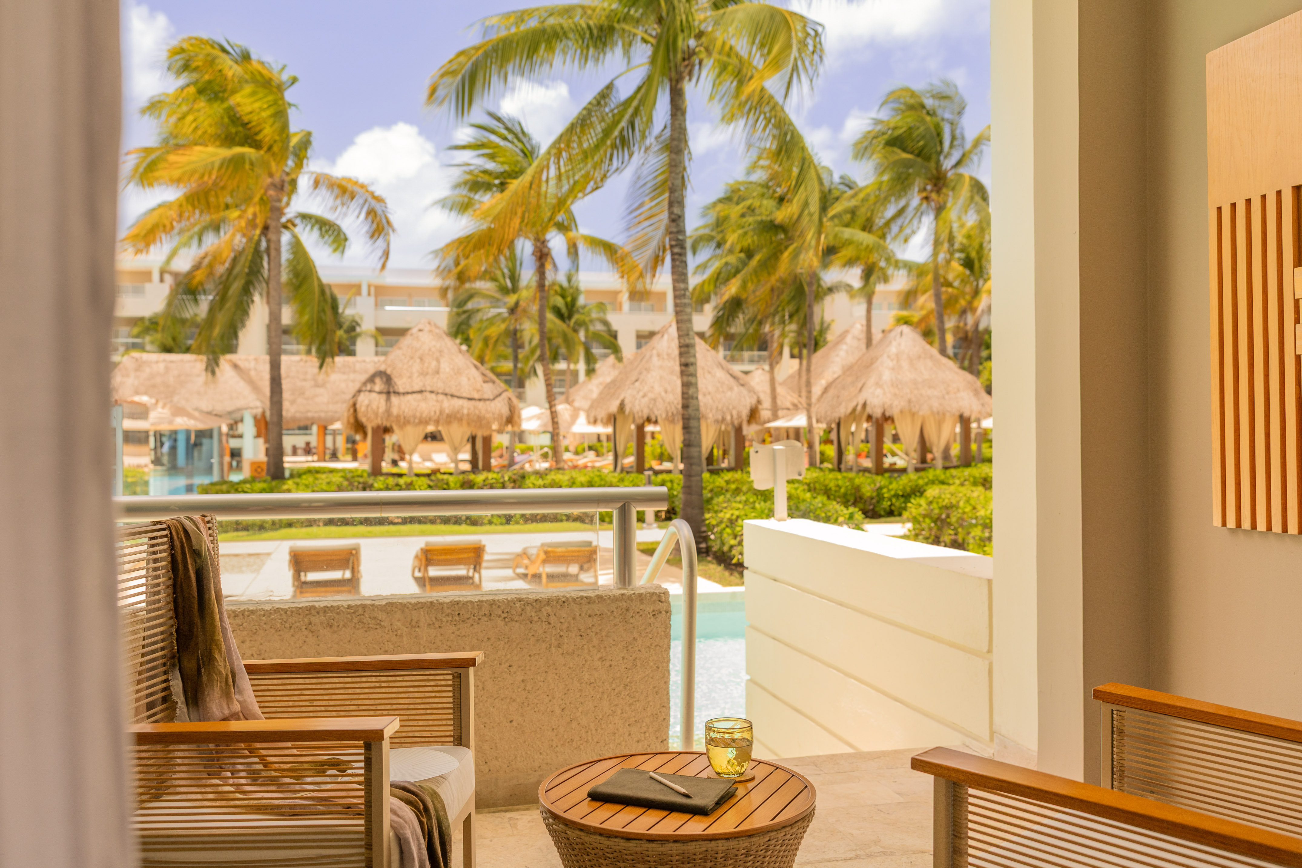 Tropical resort balcony overlooking pool with palm trees and cabanas.