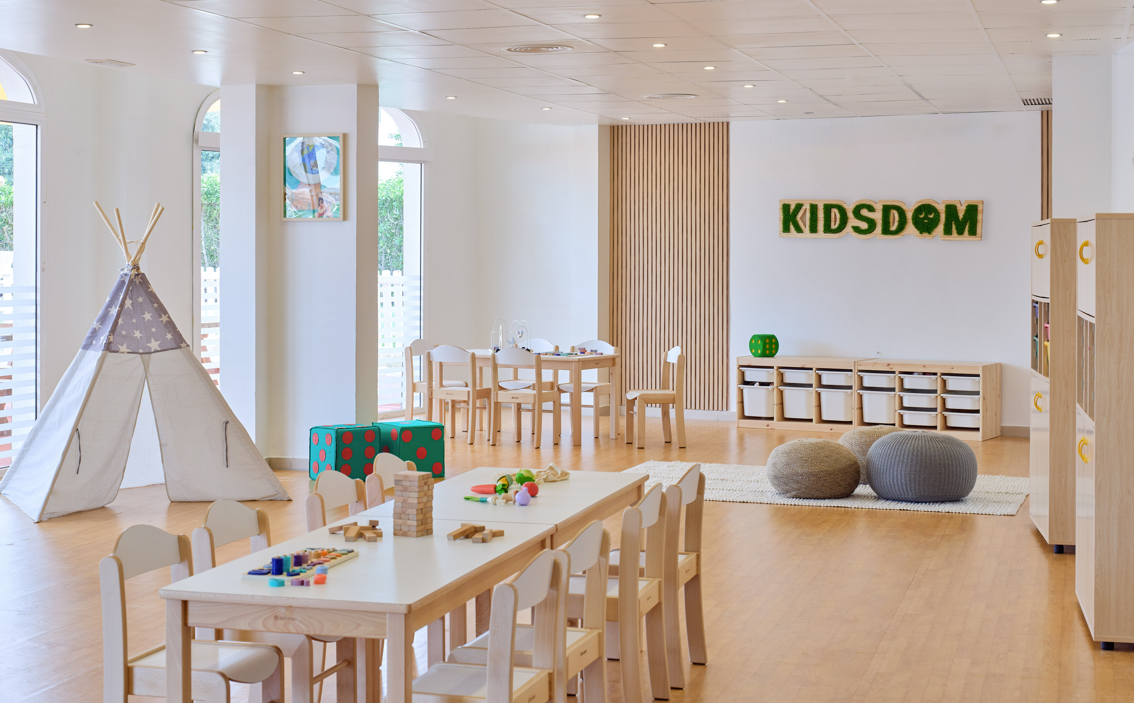 Bright kids club playroom: teepee, wooden tables, engaging toys, and "KIDSDOM" sign.