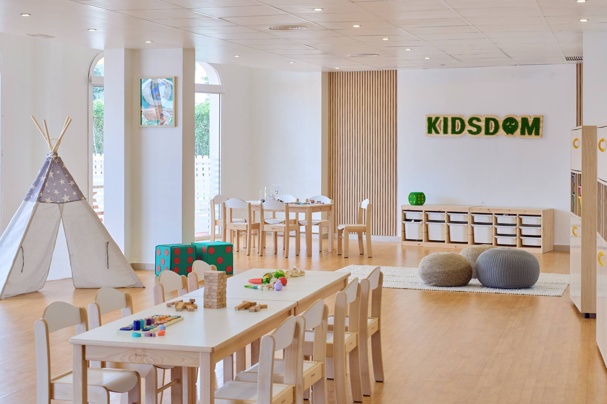 Bright kids club playroom: teepee, wooden tables, engaging toys, and "KIDSDOM" sign.