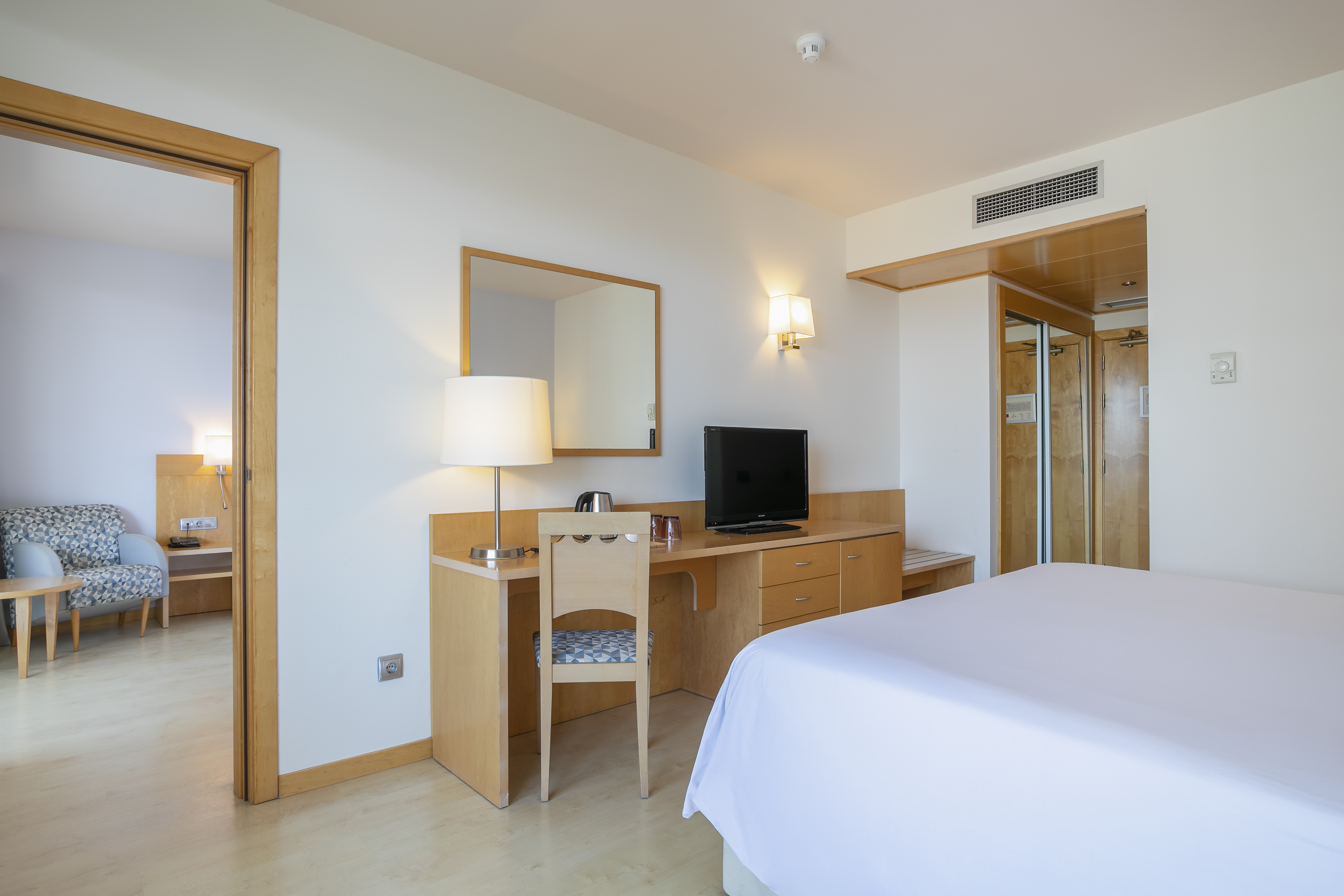 Clean hotel room with white bed, wooden desk, TV, and open adjacent area.