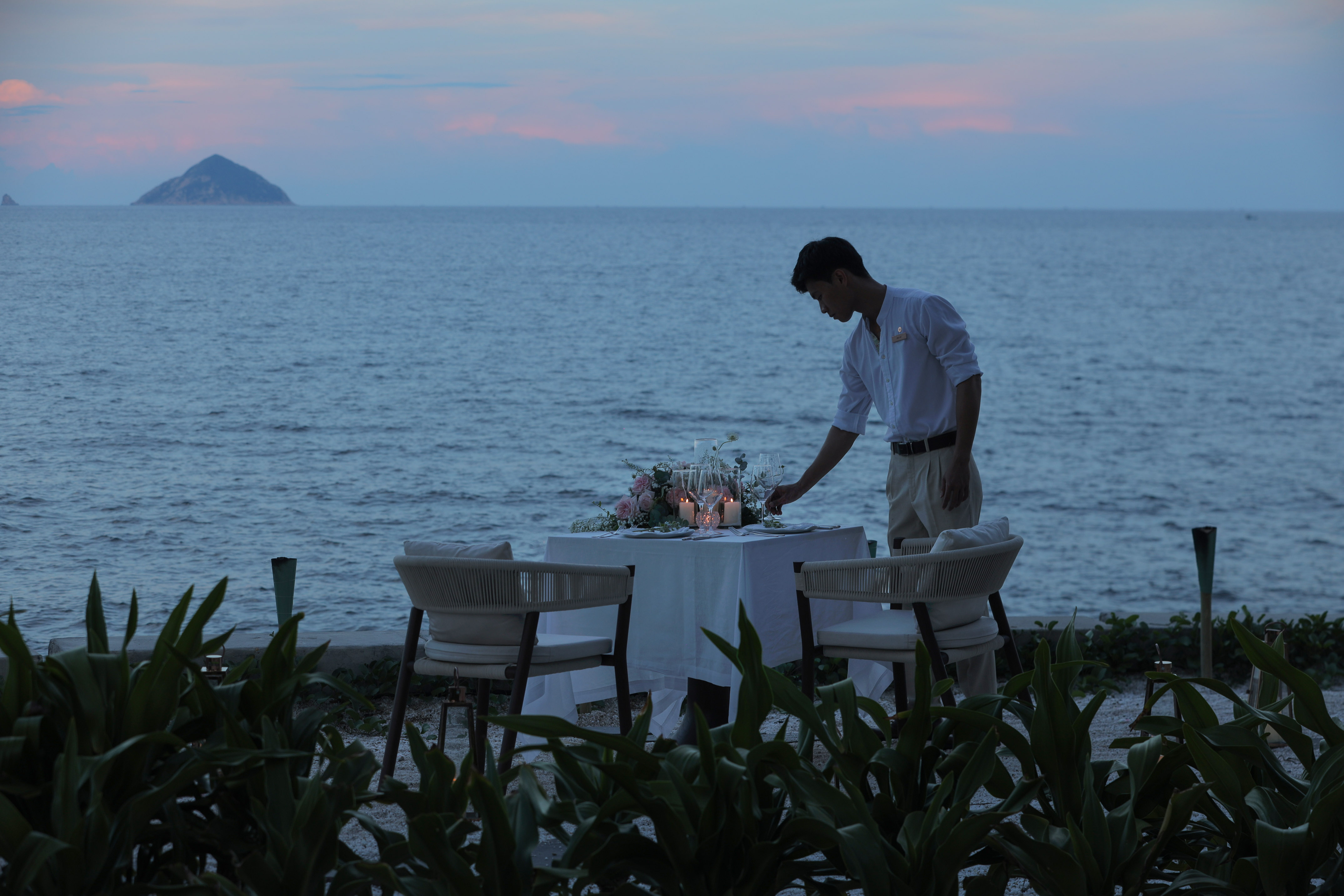 Private romantic dinner arranged by server on beach, ocean view at sunset.