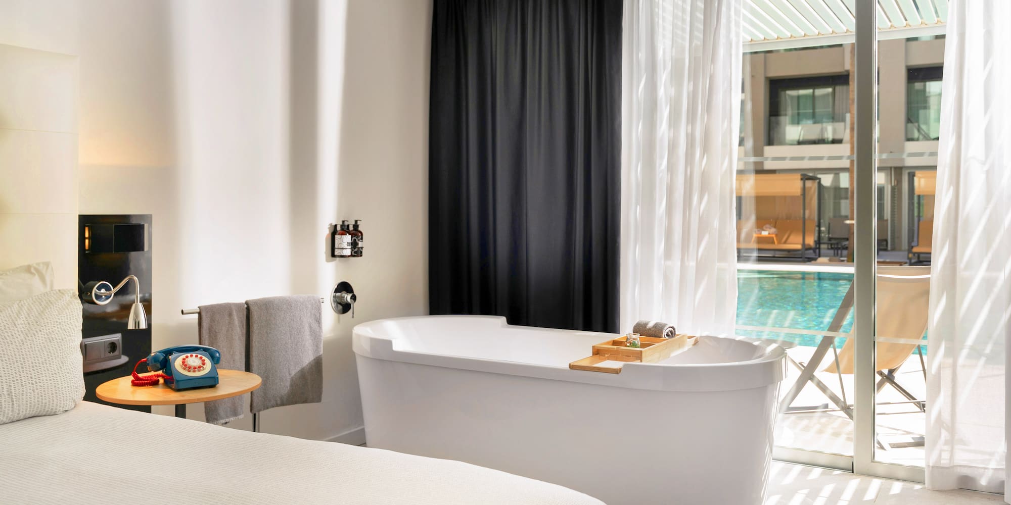 Modern hotel room with freestanding bathtub, bed, and pool view.