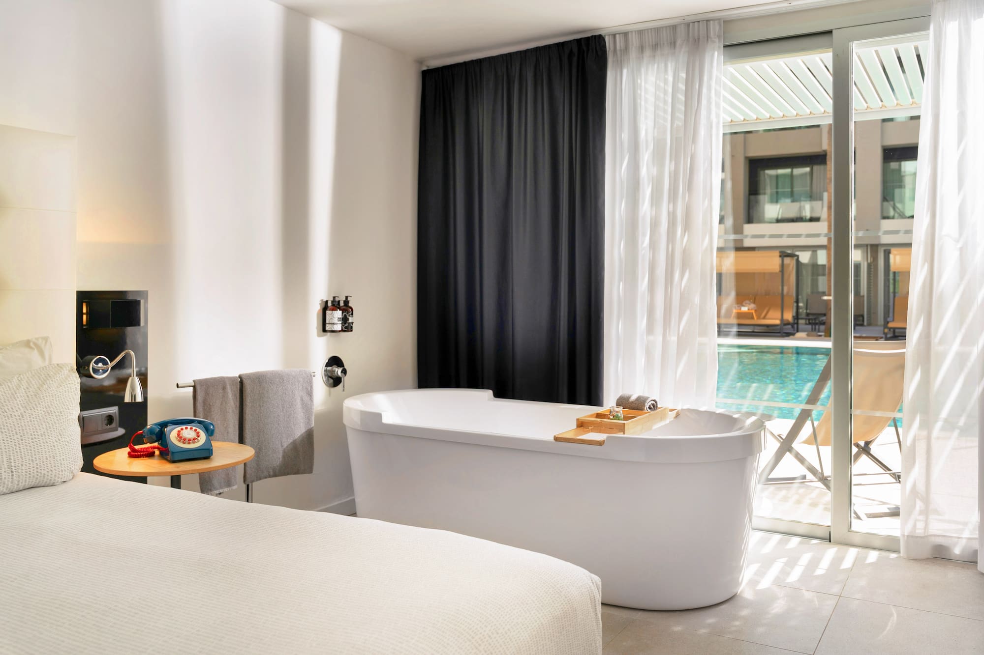 Modern hotel room with freestanding bathtub, bed, and pool view.