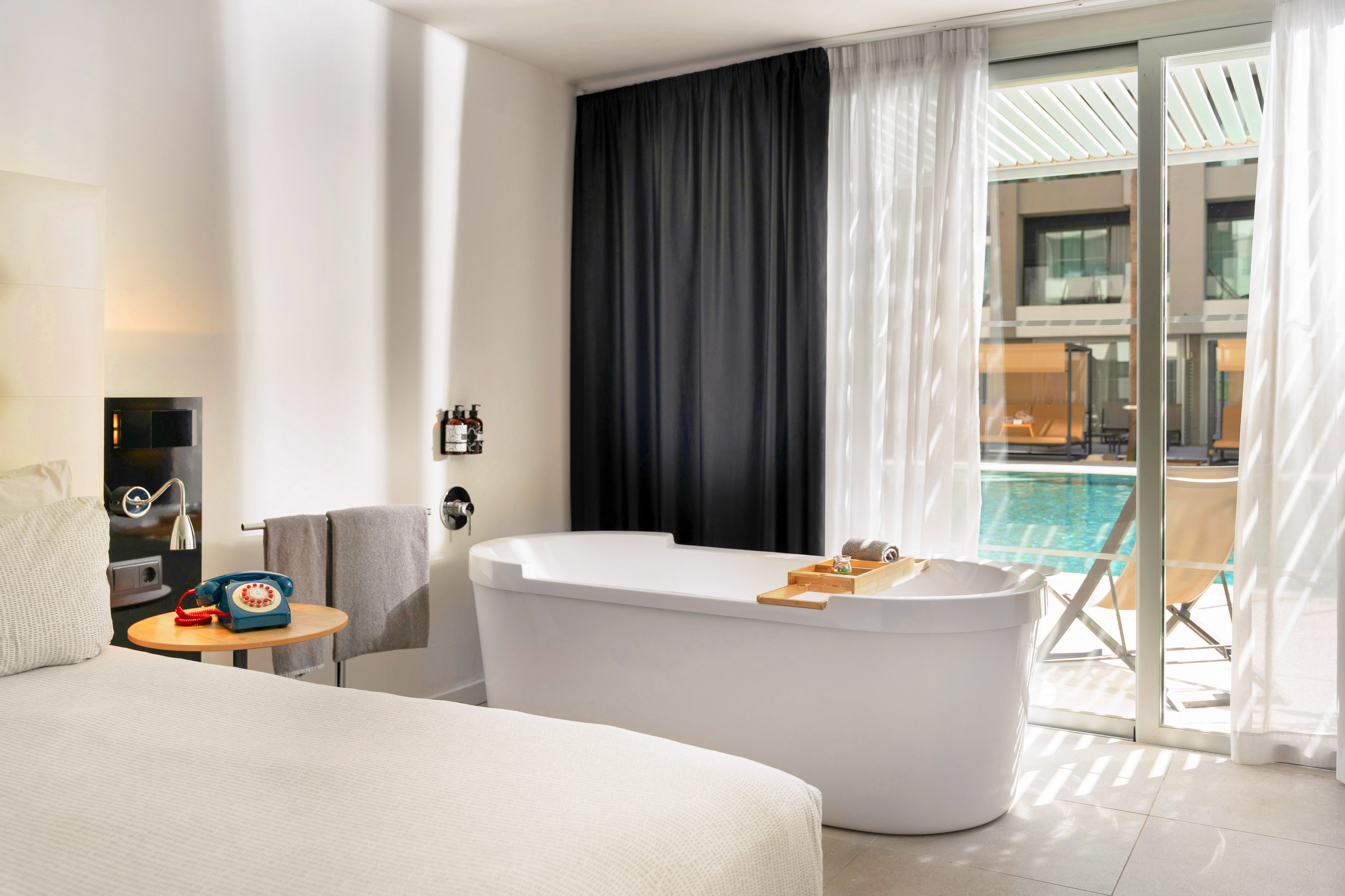 Modern hotel room with freestanding bathtub, bed, and pool view.